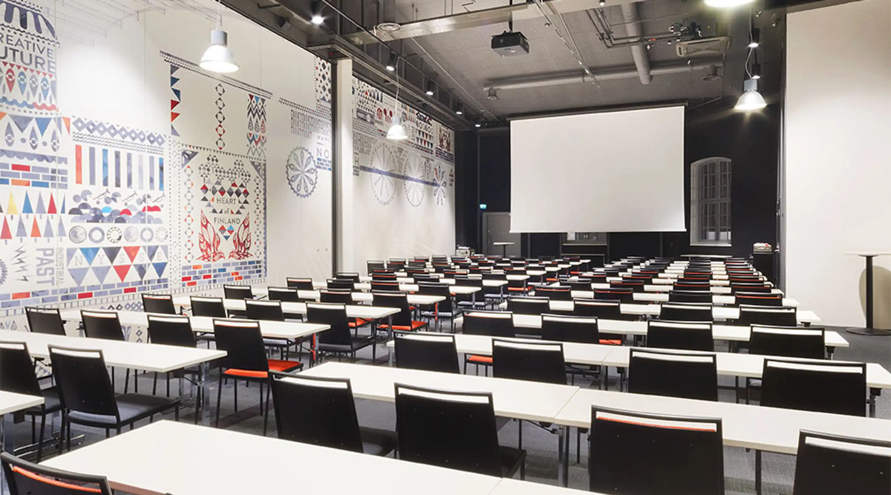 Modern auditorium with rows of tables featuring black chairs and white tabletops. Colorful geometric patterns on wall and white projection screen at front.