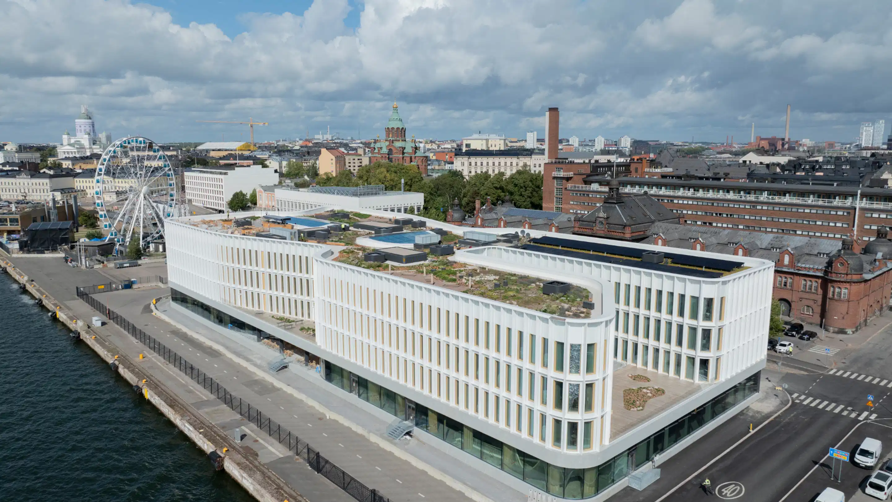 Aerial view of a modern waterfront building with green roof. Helsinki cityscape in background with ferris wheel and Helsinki Cathedral visible.