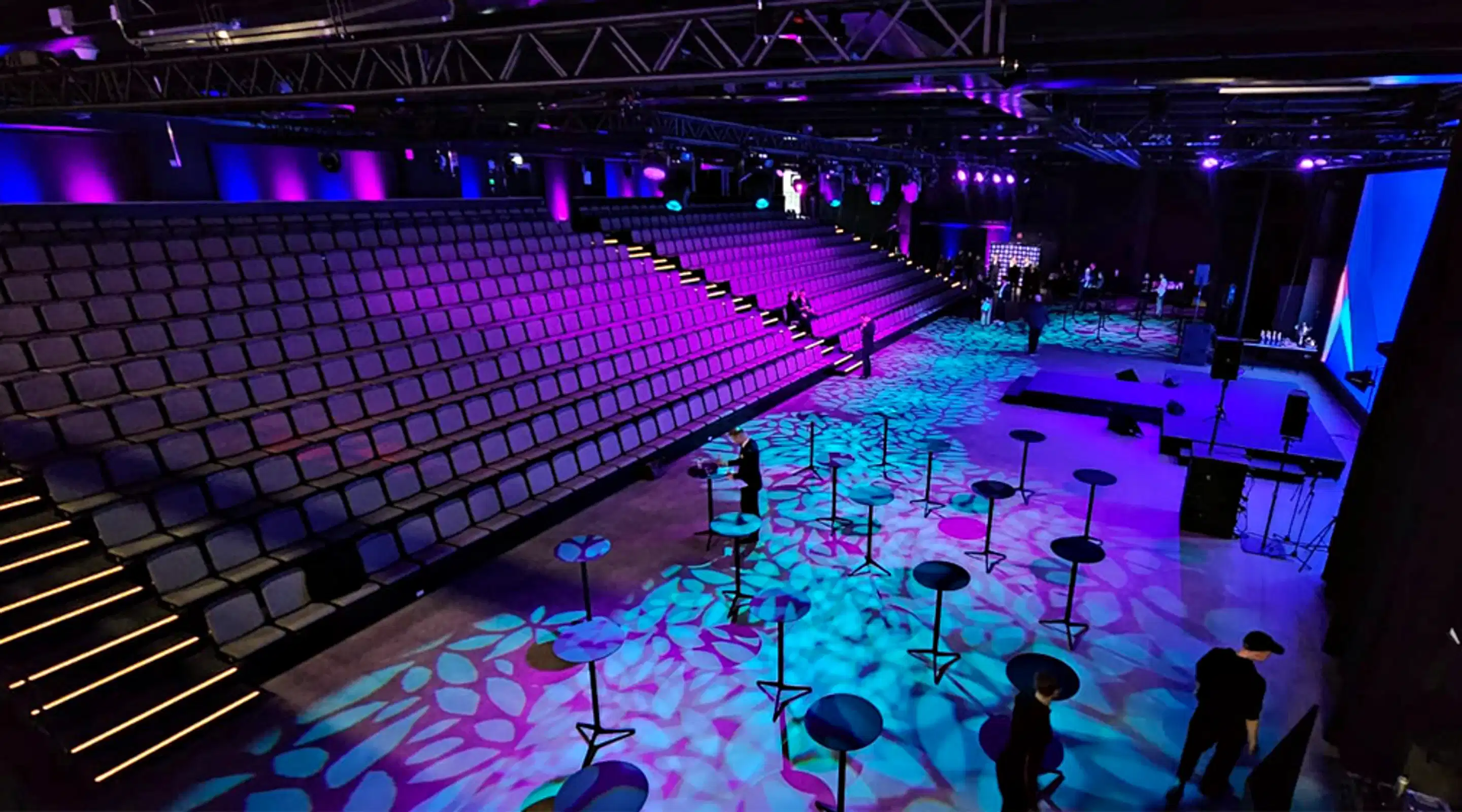 Interior of an auditorium with colorful purple and blue lighting, tiered seating on the left and stage area on the right. Standing tables on the floor with decorative light patterns.