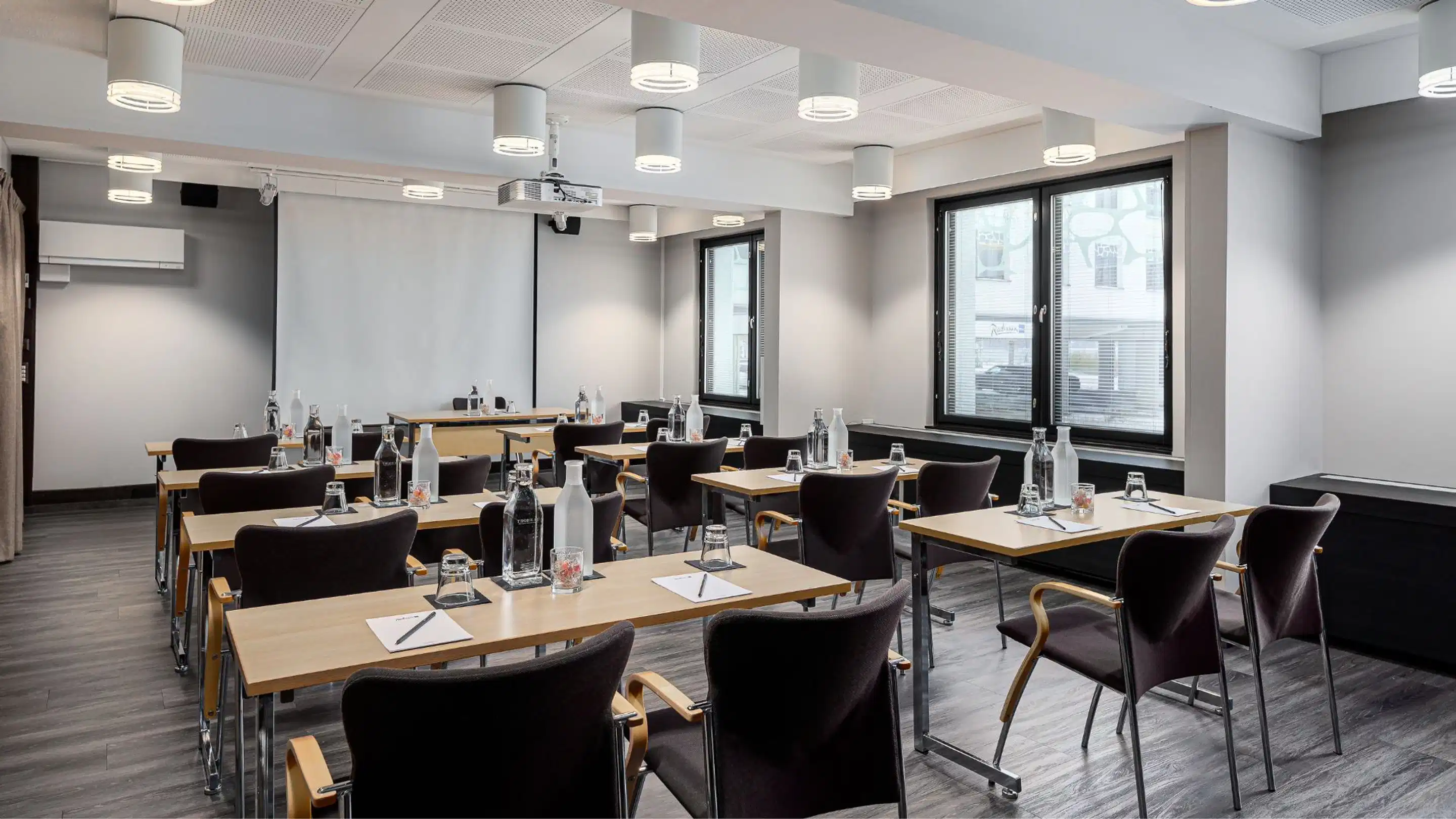 Modern meeting room with wooden tables and black chairs. The room has level flooring, large projection screen and good ceiling lighting.