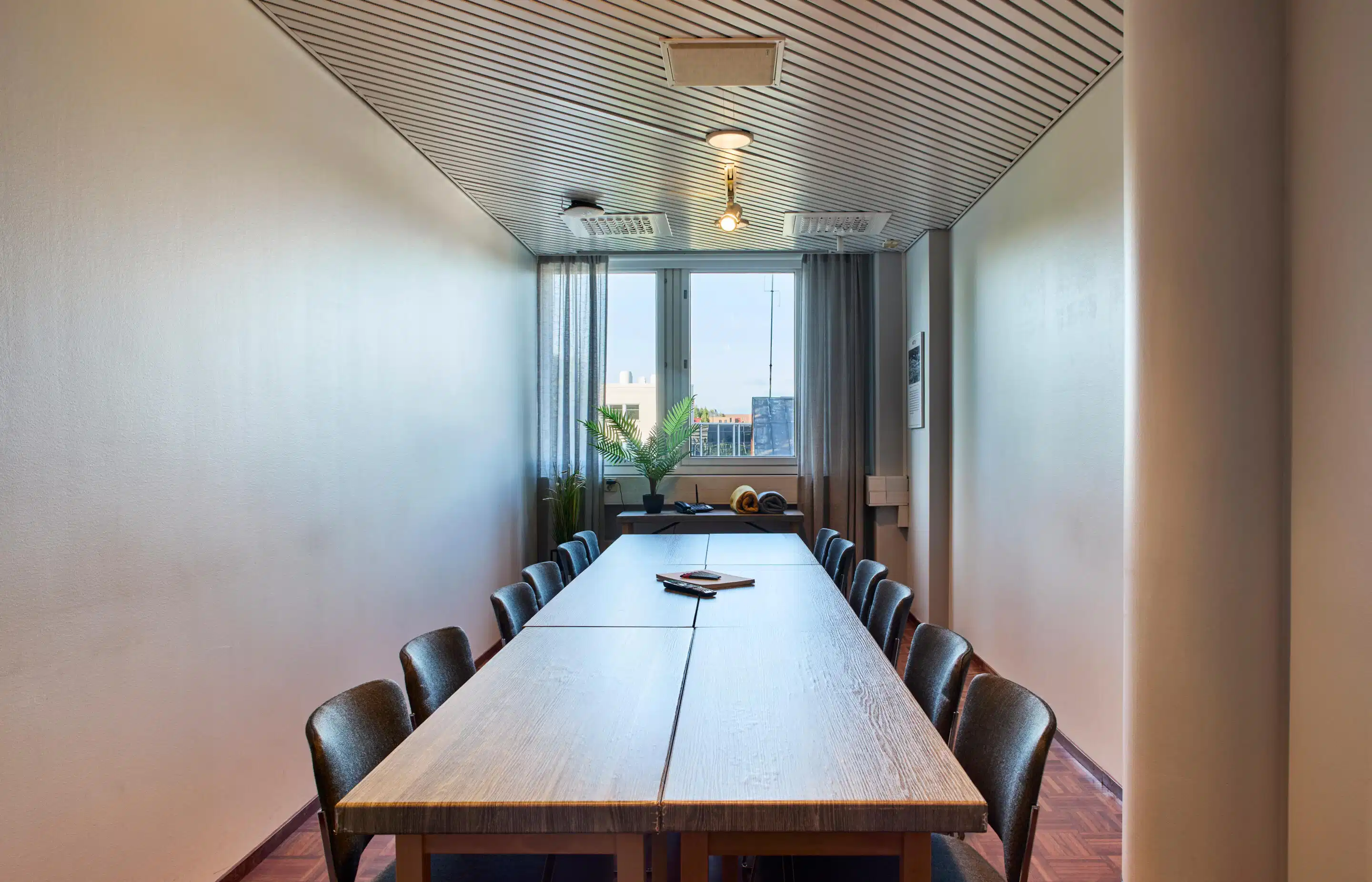 Narrow meeting room with a long wooden table and chairs. City view through the window with a potted plant on the windowsill.