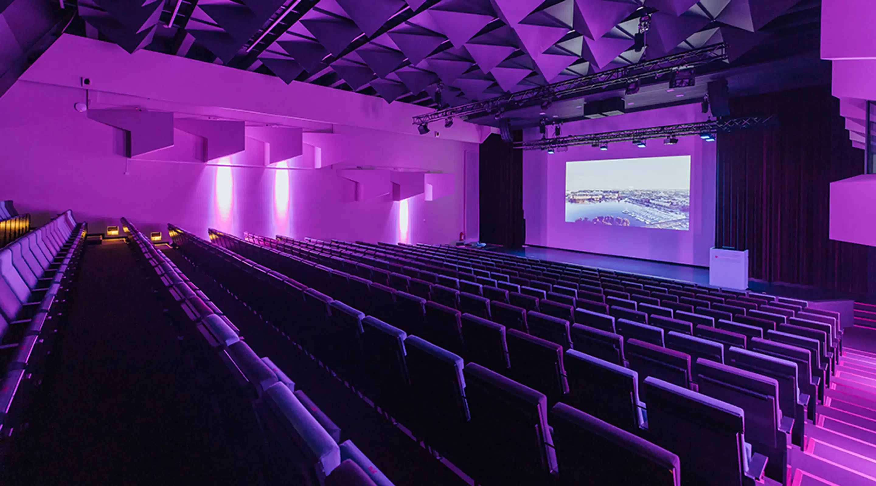 Auditorio, jossa on purppuranvärinen valaistus ja kaltevalla lattialla rivejä pehmustettuja istuimia. Edessä on valkokangas ja lava. Käytävät on valaistu himmeästi.