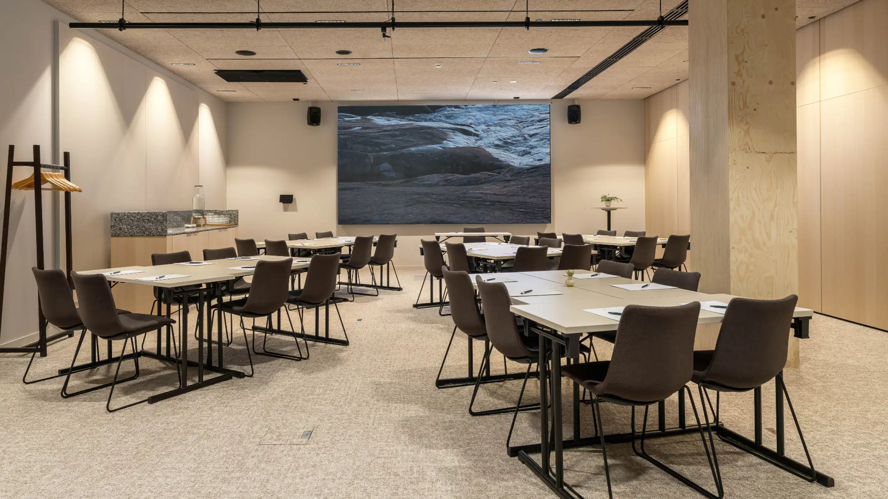 Modern meeting room with table clusters, brown chairs, large display screen at front and level carpeted floor. Light wood panels on walls and spot lighting.