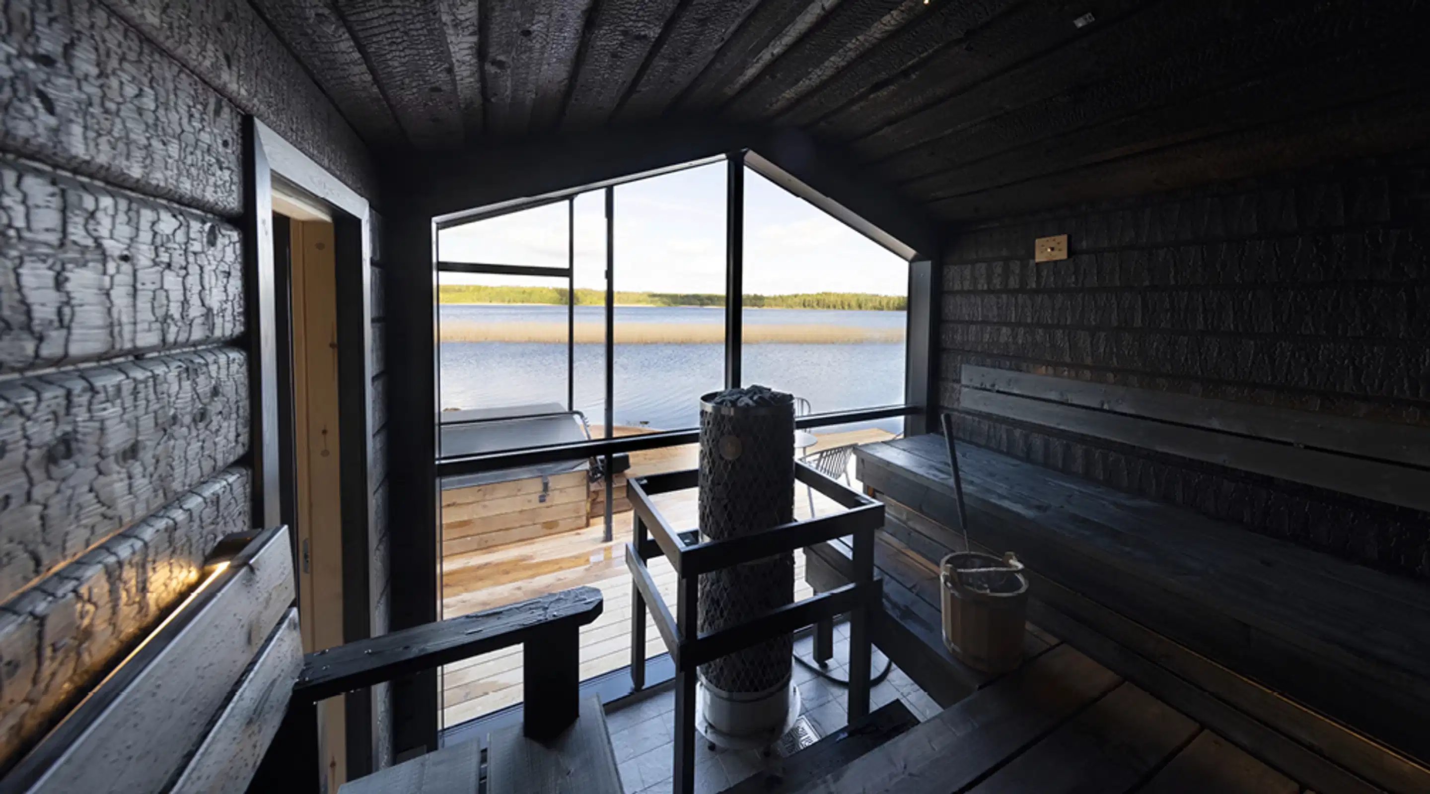 Moderni tumma sauna järvinäkymällä. Keskellä kiuas ja puiset lauteet. Suurista ikkunoista avautuu näkymä tyynelle järvelle ja metsämaisemaan.