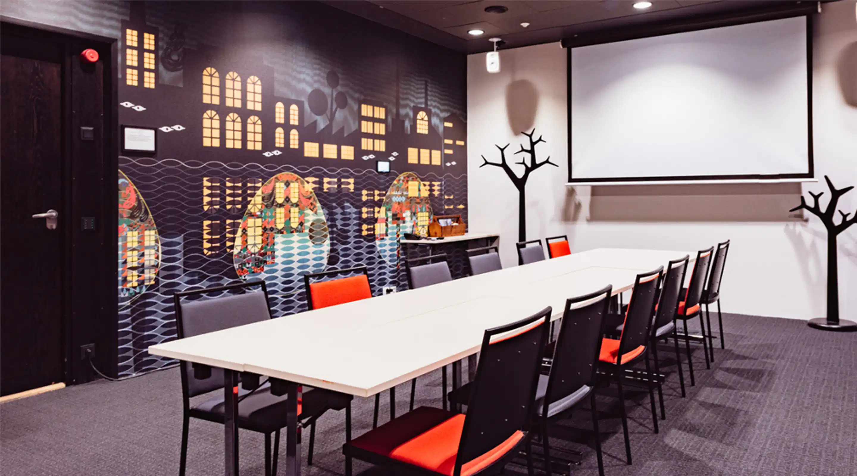 Conference room with long white table and black chairs with red seats. Wall features illustrated art design and a projector screen.