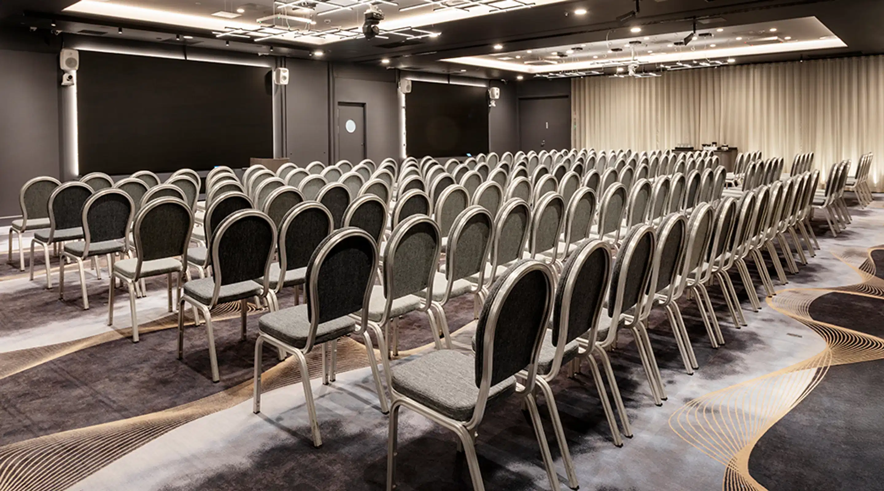 Auditorio, jossa on useita tuoliriviä kohti valkokangasta. Lattialla on kuviollinen kokolattiamatto. Tuolirivien välissä on käytäviä. Huoneessa on kohdevalaistus.