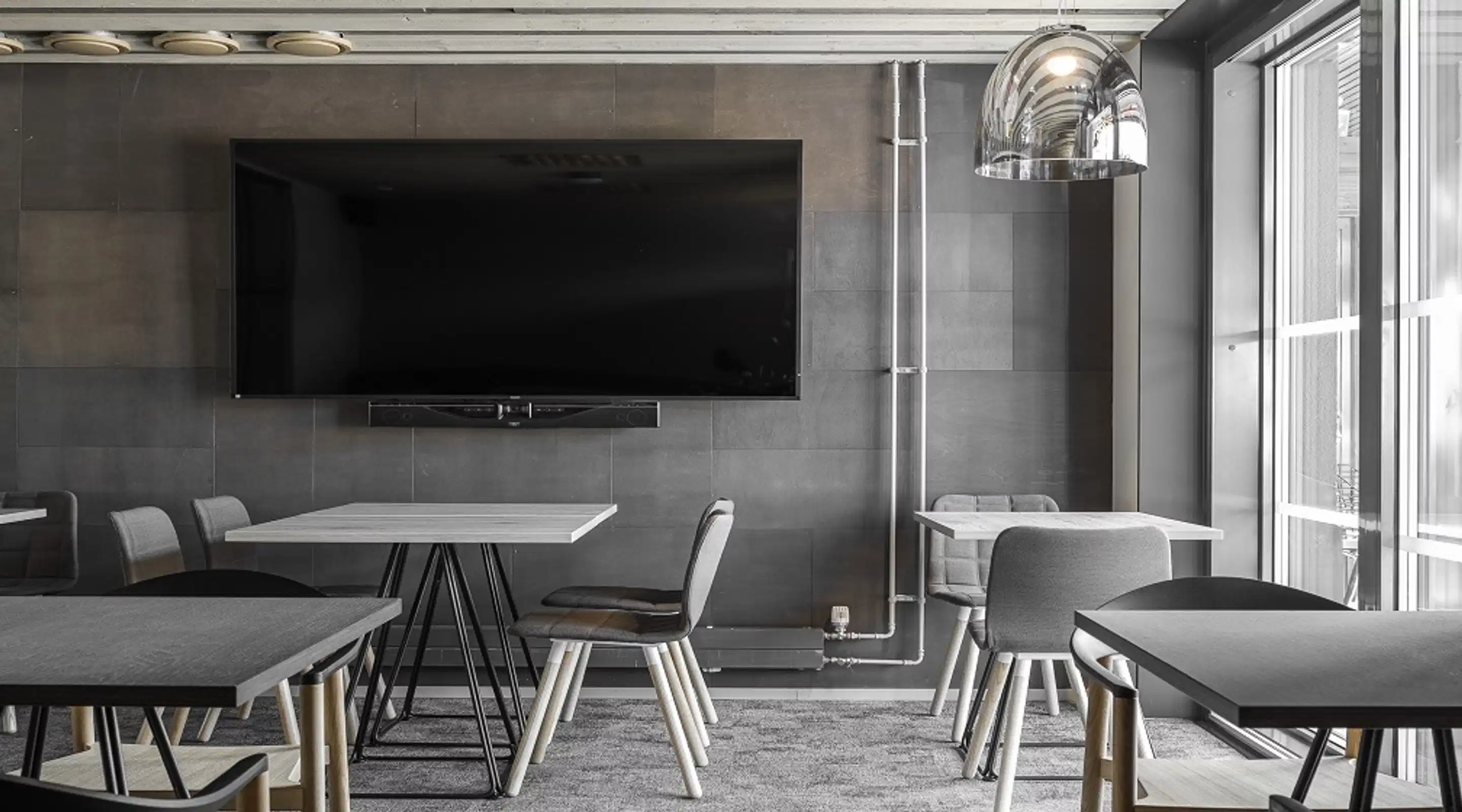 Modern meeting room with gray tables and chairs, large wall-mounted TV, and large windows allowing natural light. Hard flooring with area carpet.