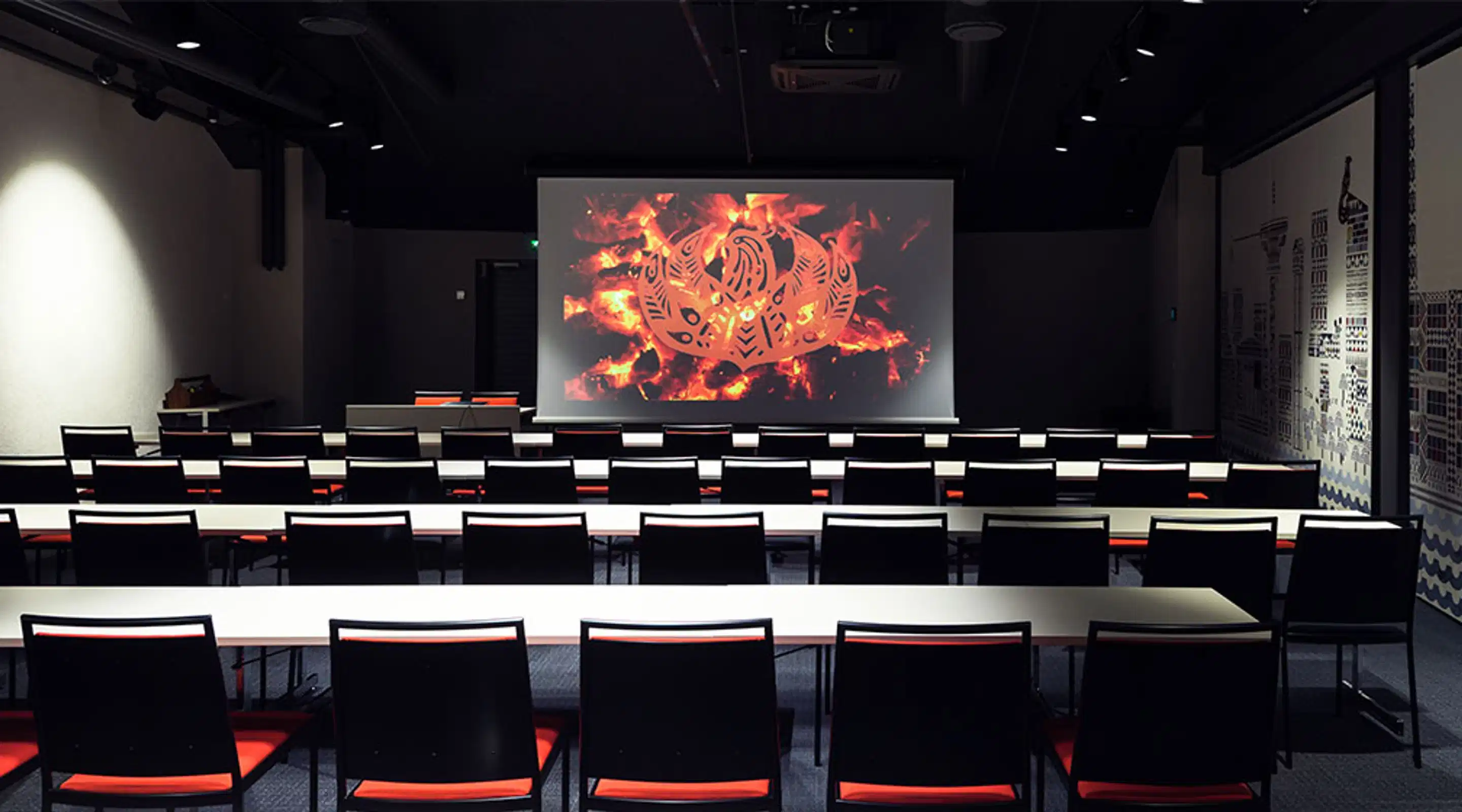 Auditorium interior with long tables and chairs. A large screen on the wall displays a fiery pattern. The room is dark except for the projector-illuminated screen.