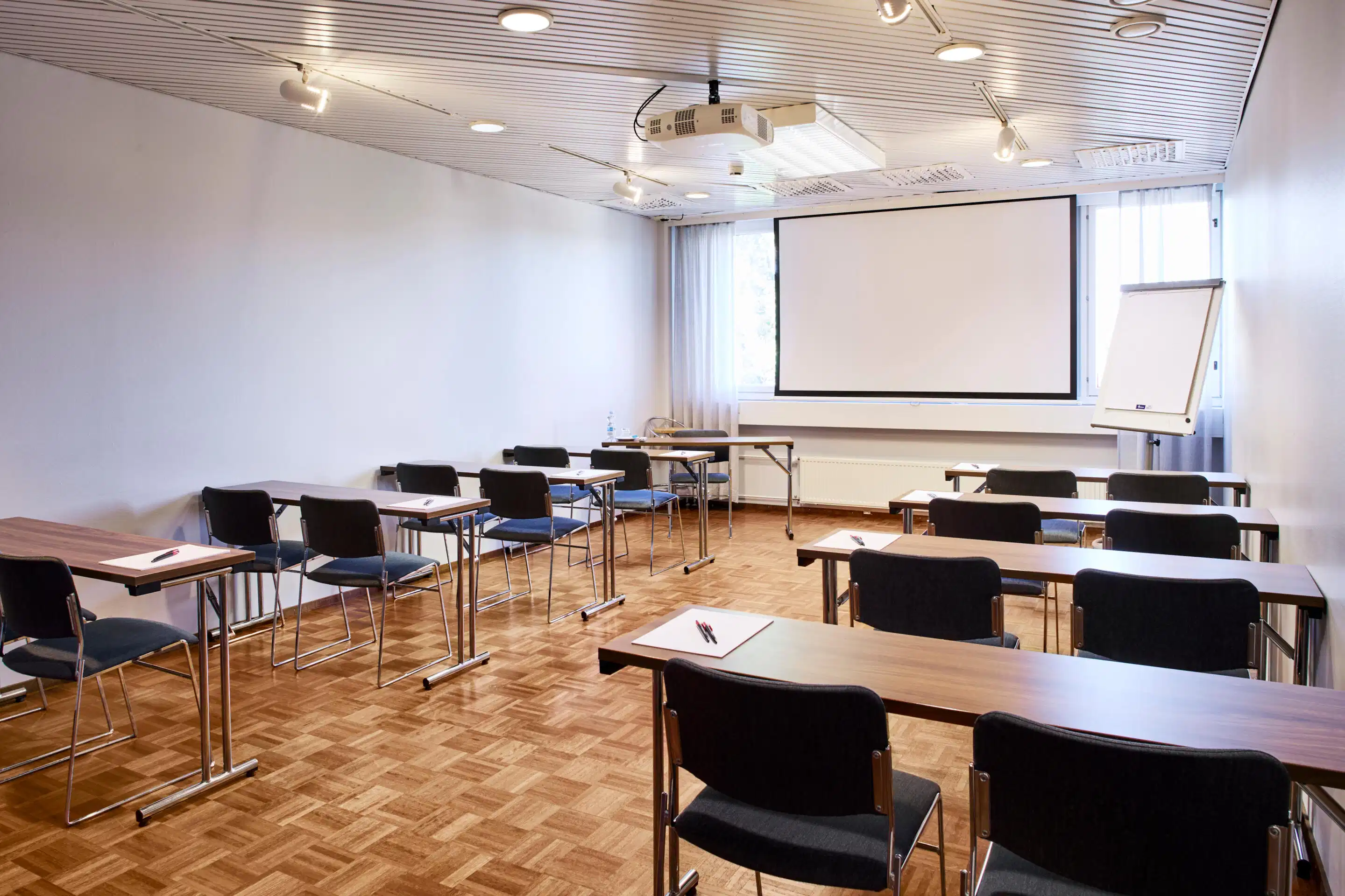 Training room with multiple tables and chairs, projection screen at front, parquet flooring and good ceiling lighting.