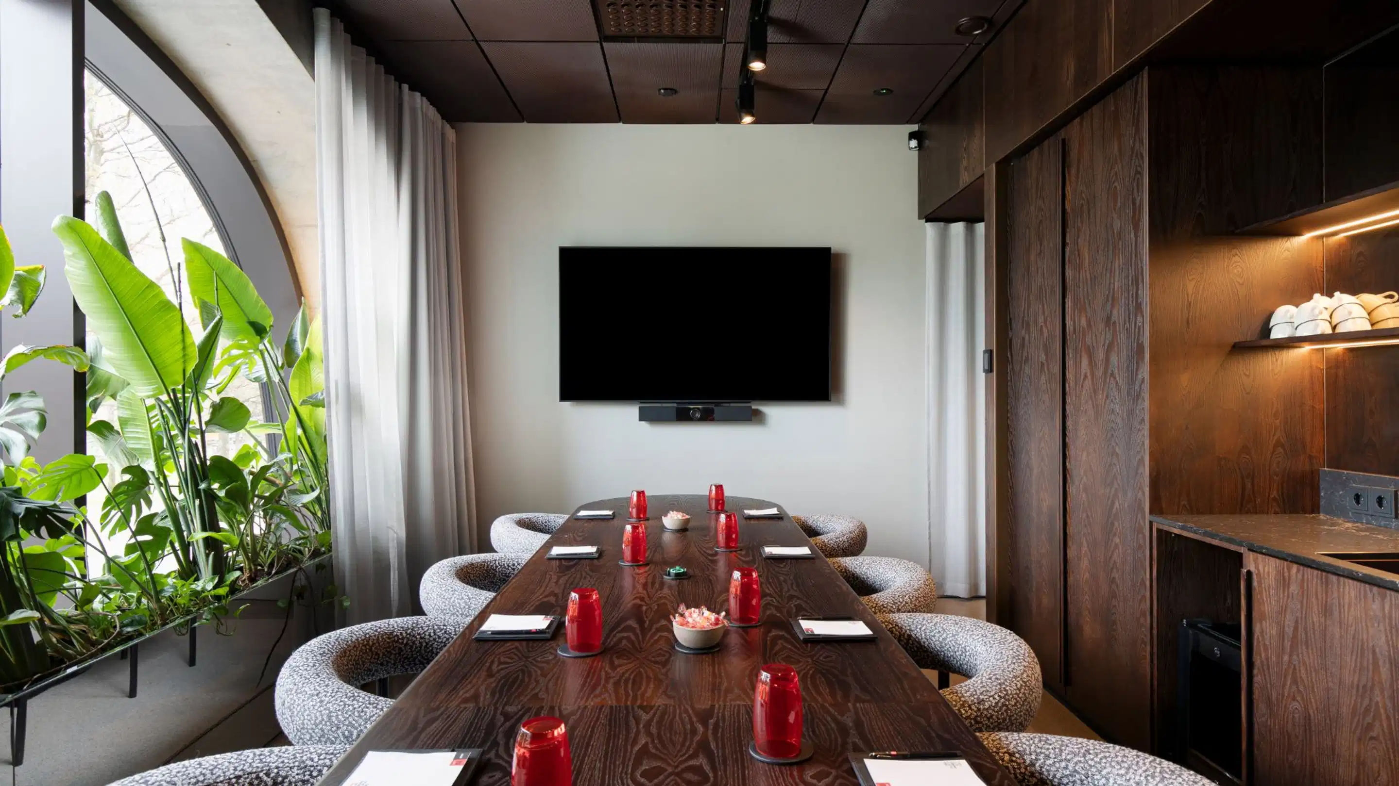 Modern meeting room with wooden table, comfortable chairs and wall-mounted screen. Large arched window brings natural light into the space.