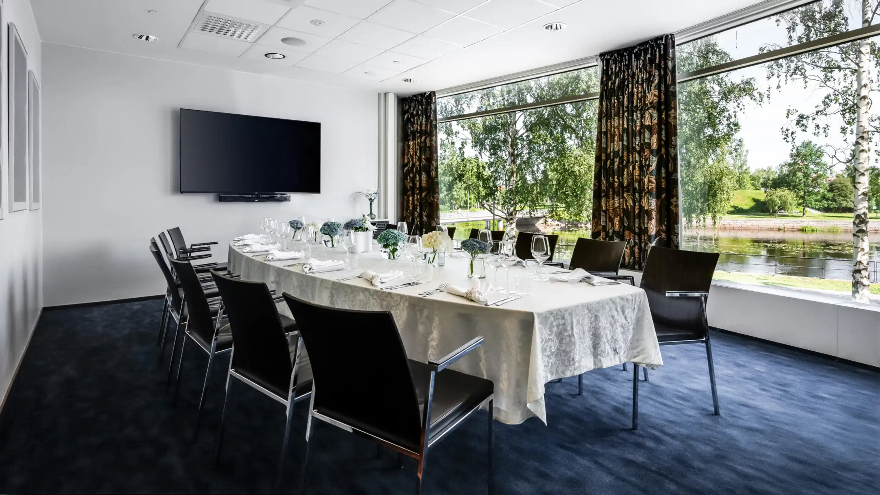 Meeting room with a set table, large windows overlooking a lake view and a wall-mounted large TV. Level floor with blue carpeting.