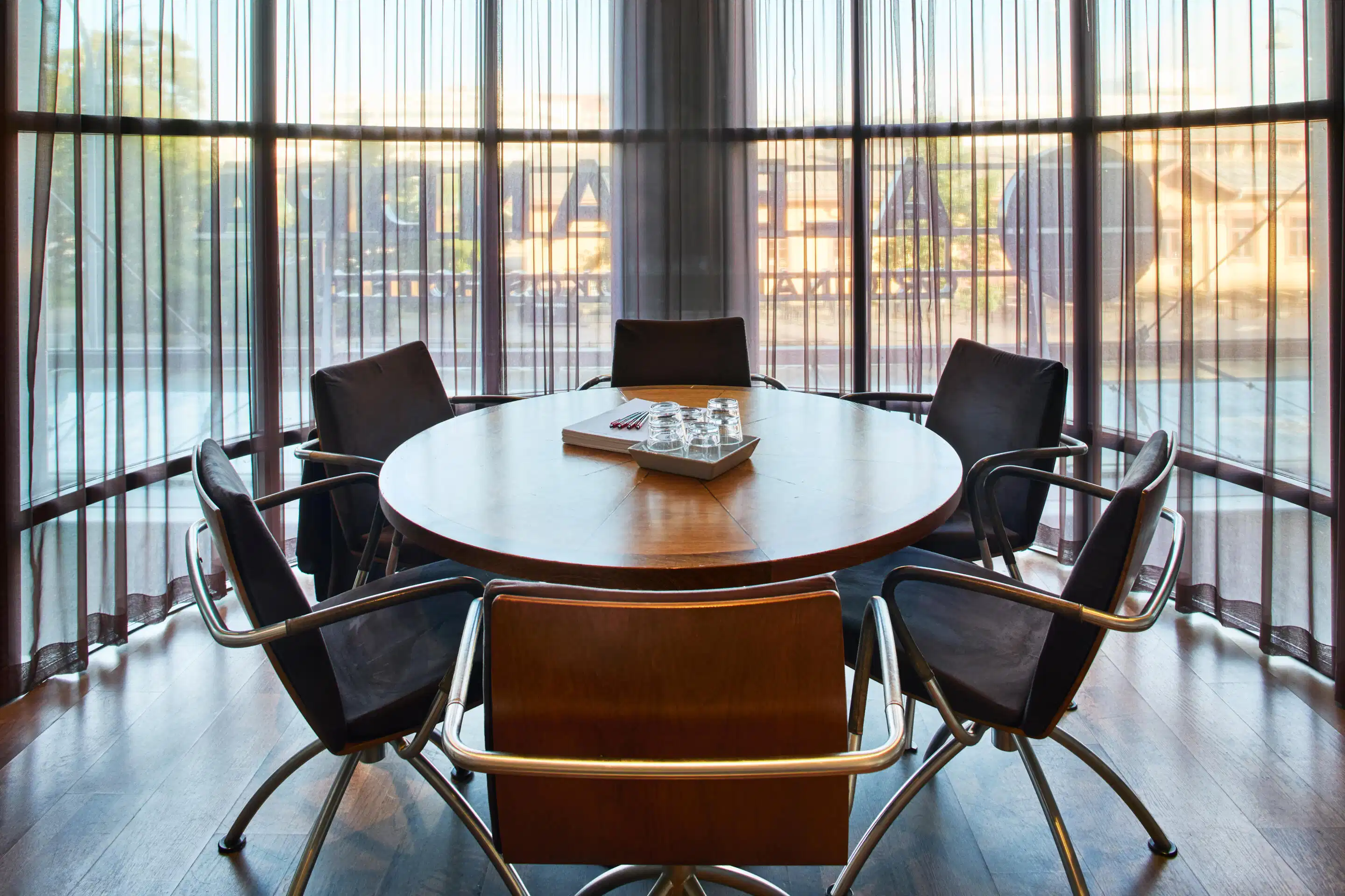 Round conference table with six chairs in front of glass facade. Wooden floor and view of buildings through sheer curtains.