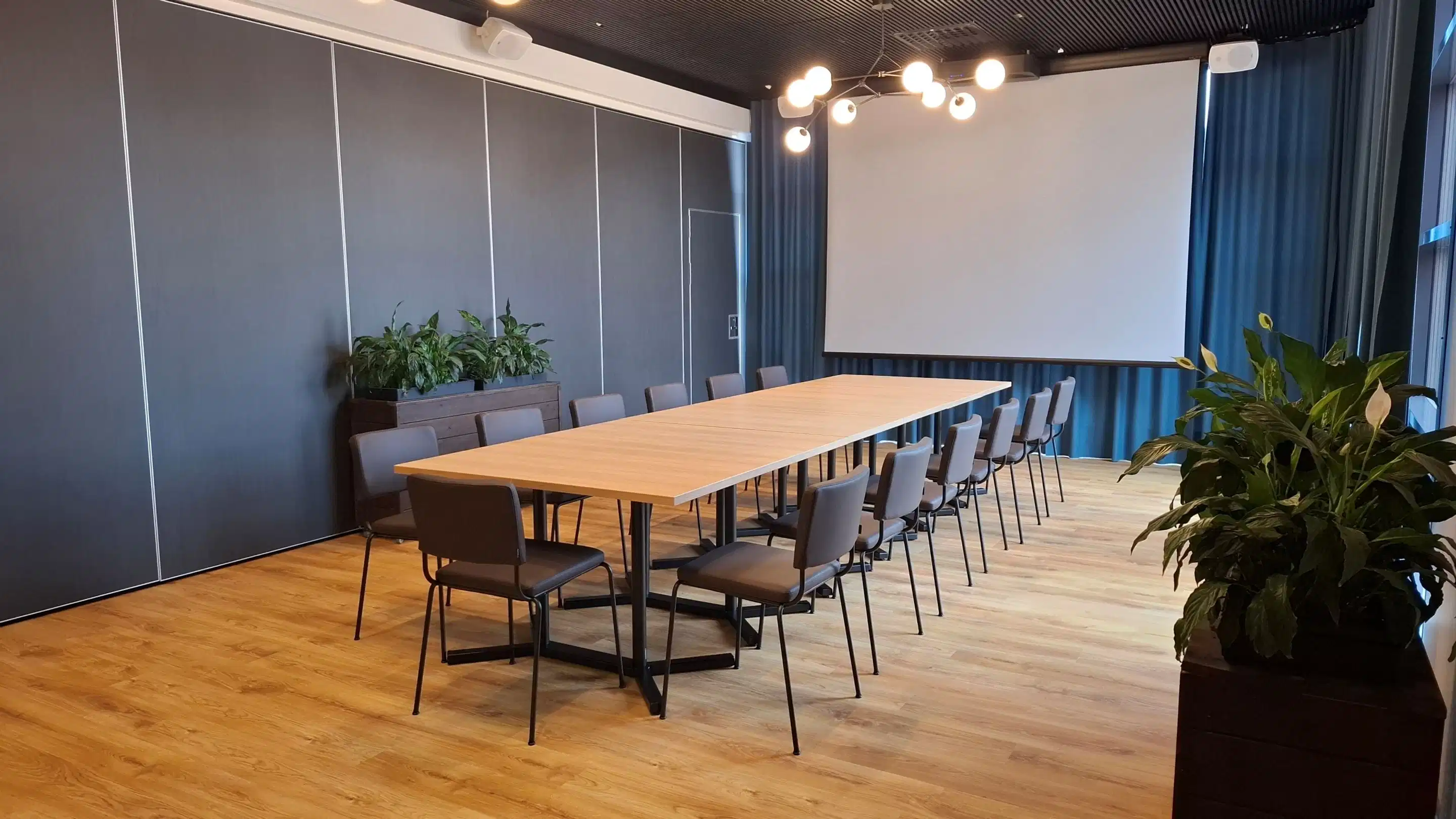 Modernt mötesrum med långt träbord, stolar på båda sidor, vit projektorduk och gröna växter. Jämnt trägolv.