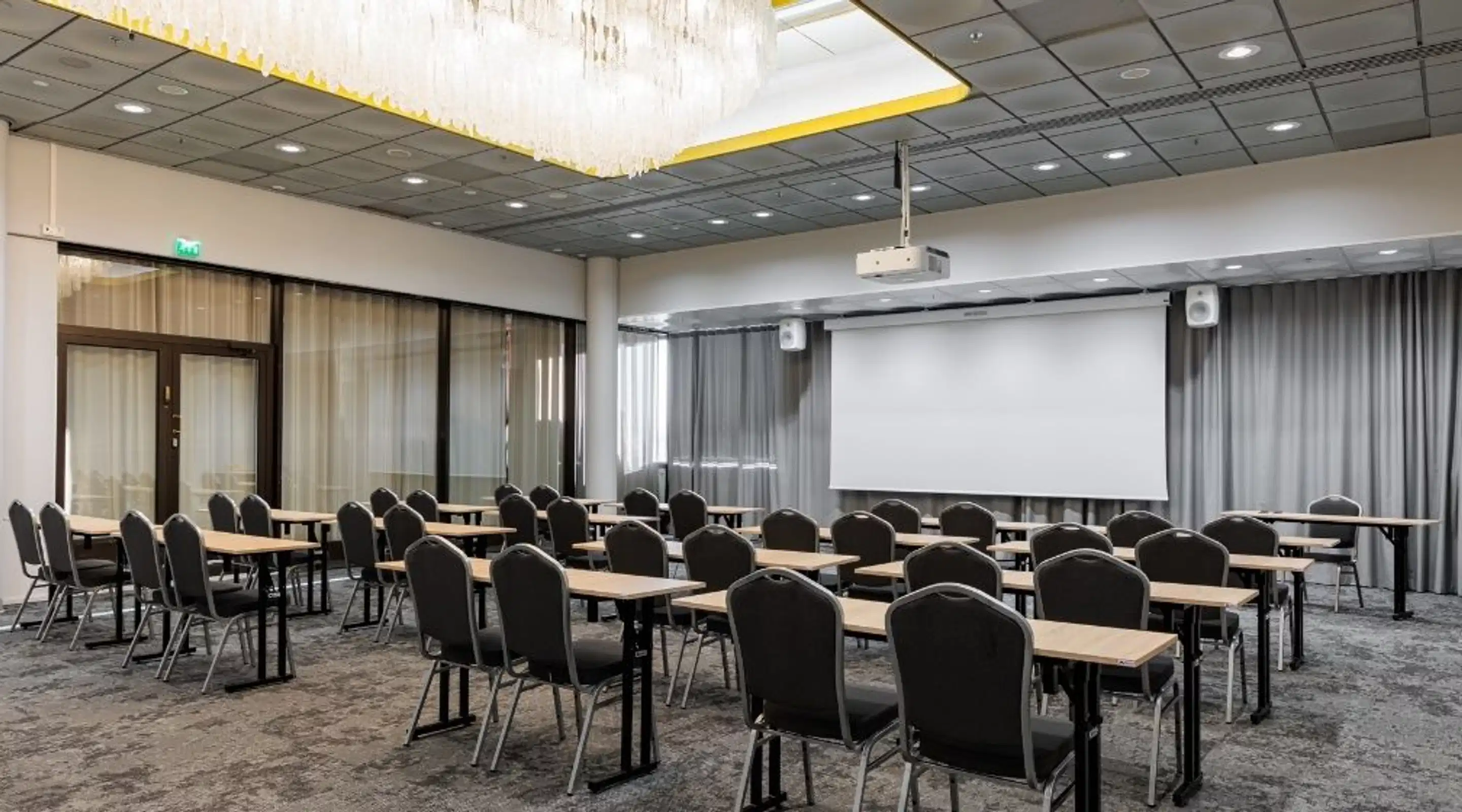 Conference room with rows of tables and chairs, large chandelier overhead, projector screen at front. Room features large windows with grey curtains.