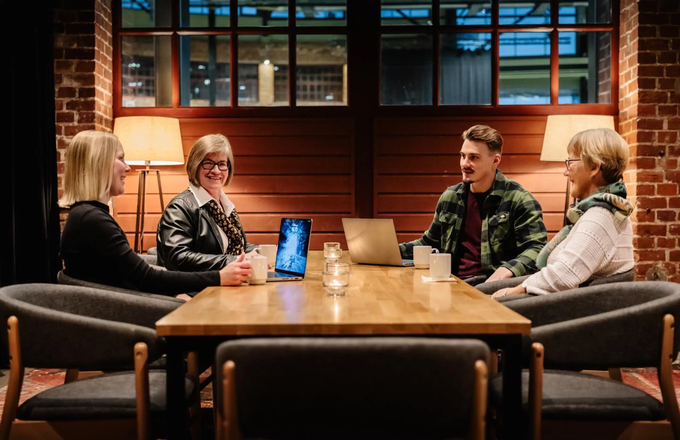 Grupp inimesi puidust laua ümber modernses ruumis, millel on tellisseinad ja soojad lambid. Laual on sülearvutid ja joogid.