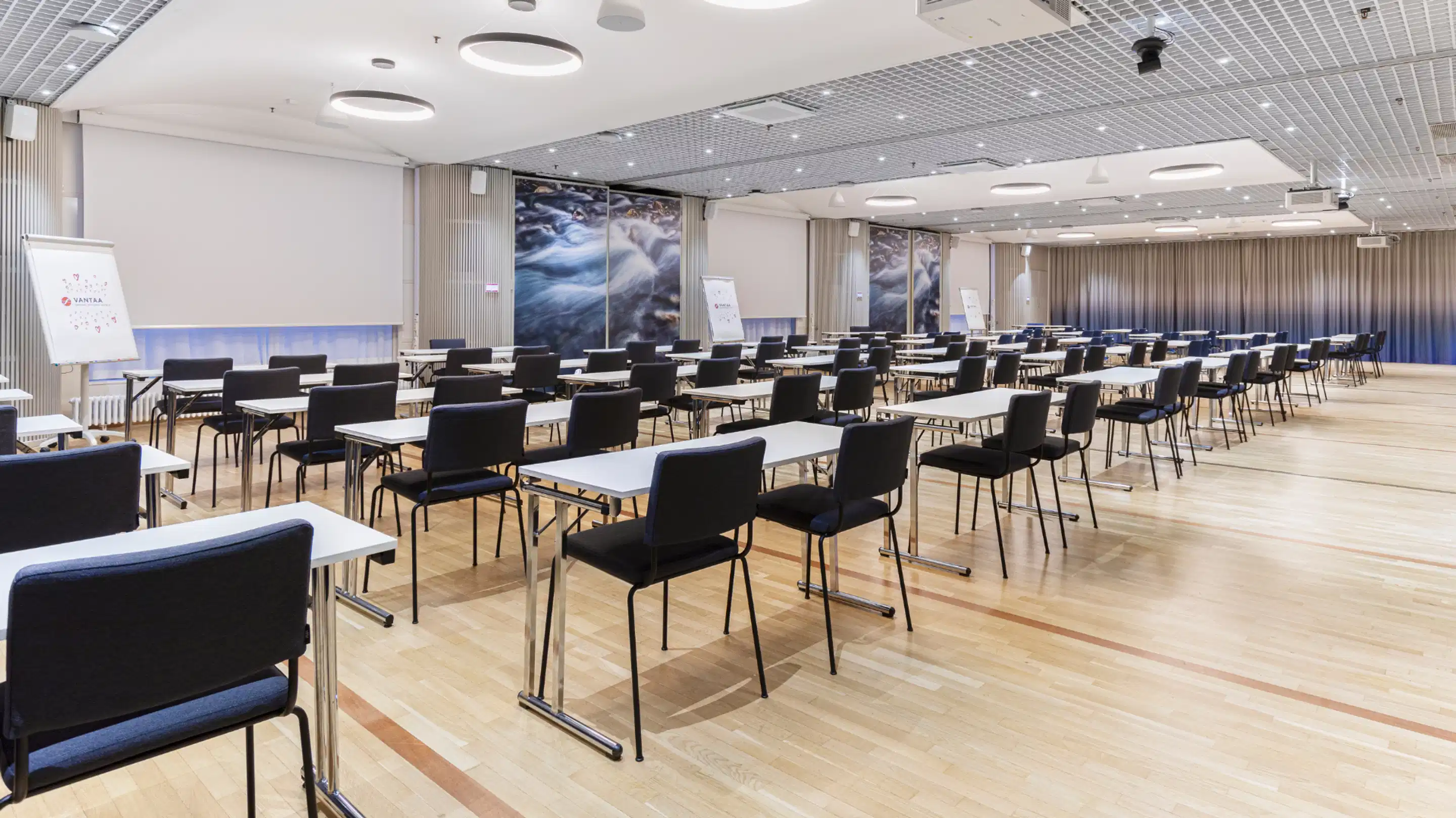Modern conference room with bright wooden floor, black chairs and white tables arranged in rows. Nature-themed wall art and flipcharts visible along the sides.