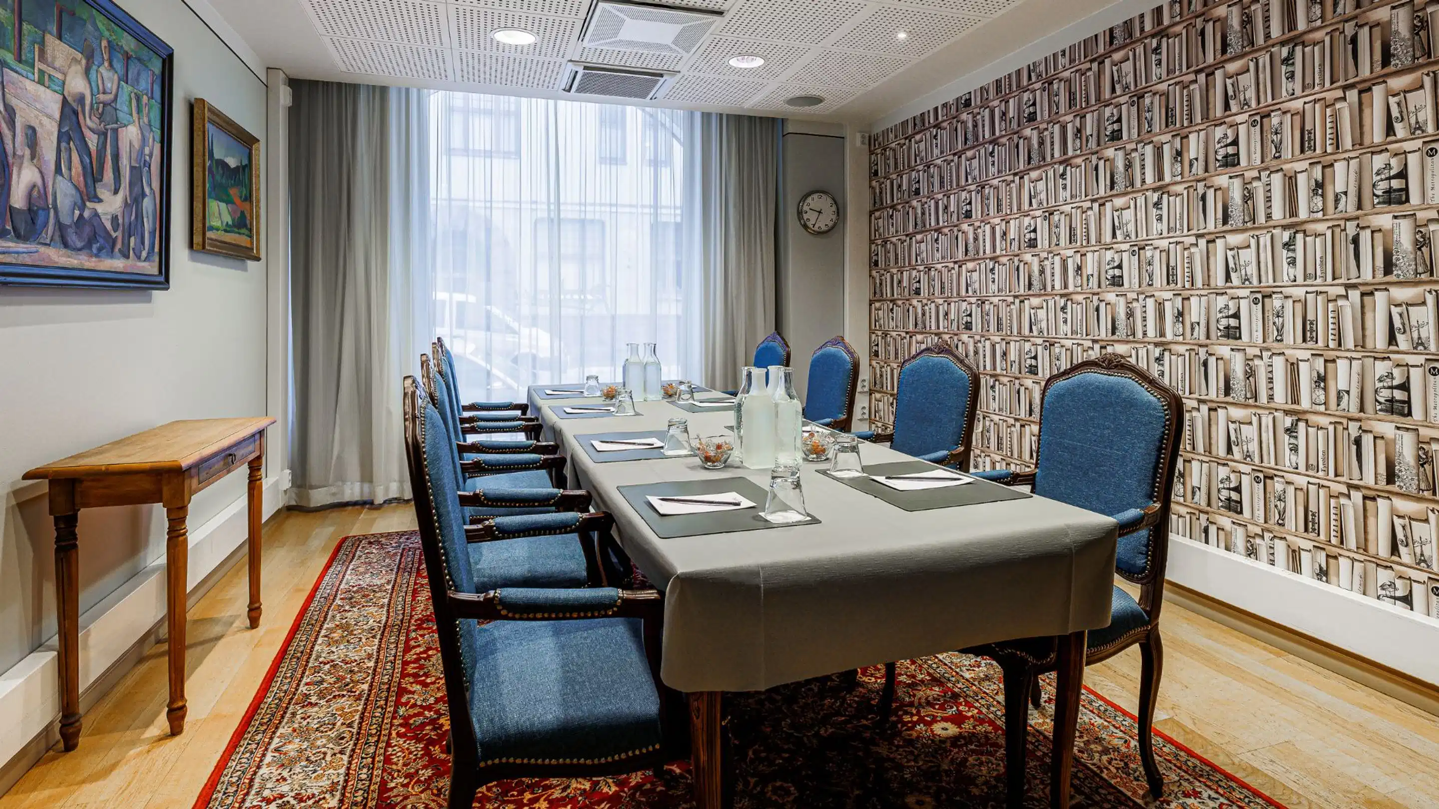 Meeting room with bookshelf wallpaper, long table with blue chairs, wooden floor, windows and artwork on the wall.
