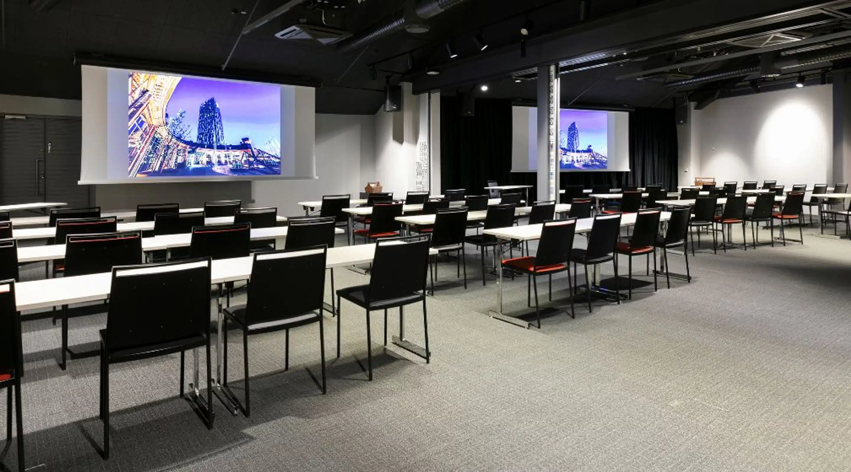 Auditorium with rows of white tables and black chairs on flat carpet. Two large projection screens on walls.