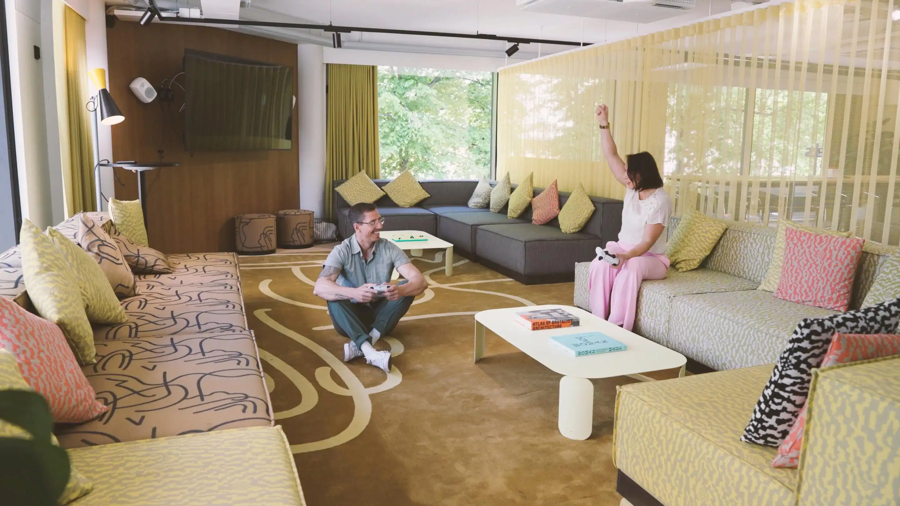 Modern lounge area with multiple sofas and colorful cushions. Spacious room with large windows, patterned carpet and two people playing video games.