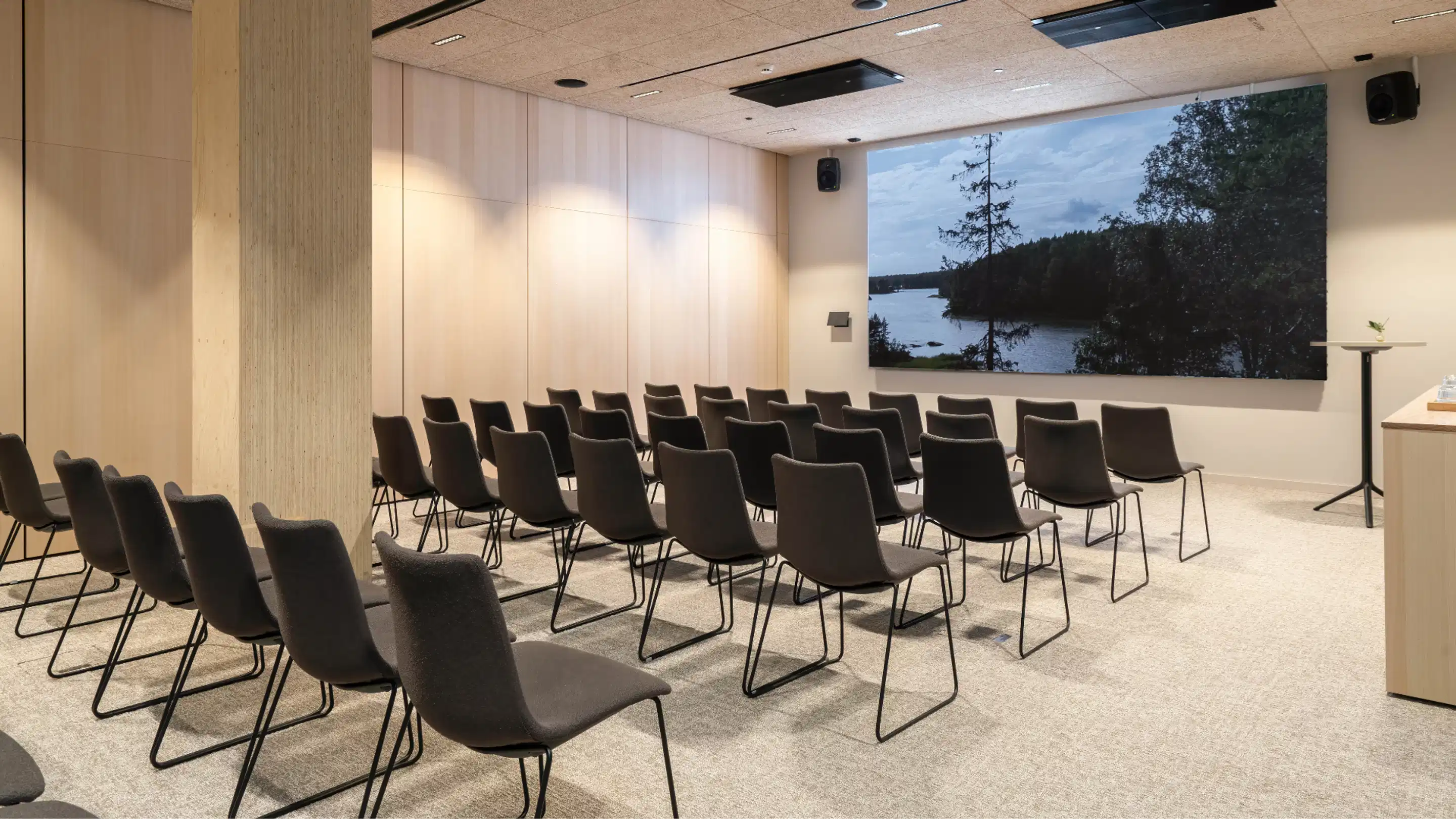 Modern auditorium with chairs arranged in rows. Large nature image of a lake landscape on the wall. Light flooring and wooden panel walls.