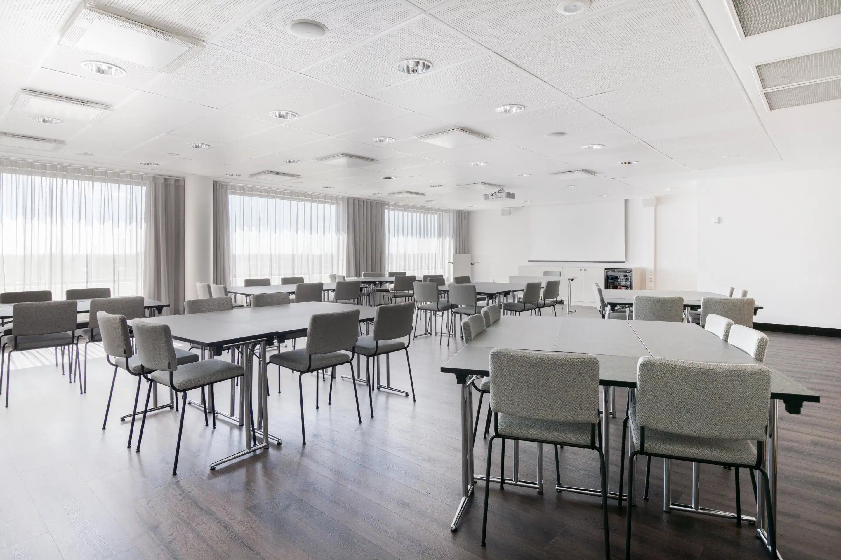 Bright meeting room with grey chairs and rectangular tables. Smooth wooden floor and large windows. Projection screen on wall for presentations.