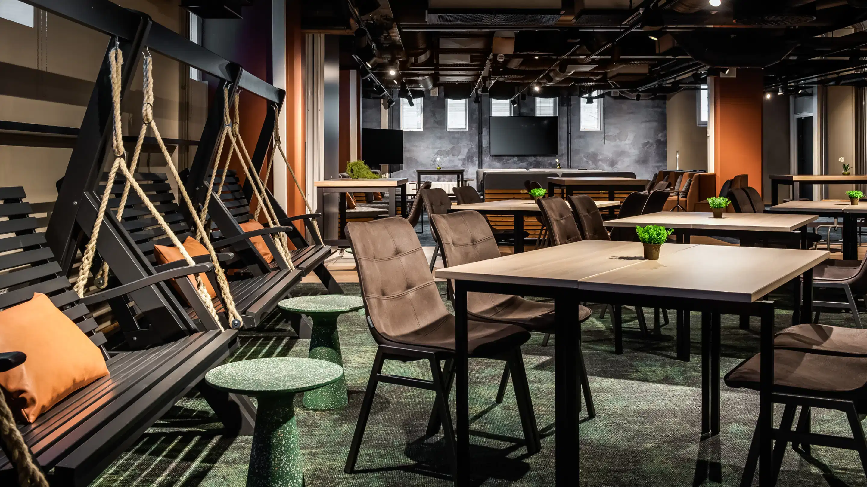 Modern multi-purpose office space with tables, swing chairs hanging from ropes, and green carpeting. The space features screens and small potted plants on tables.