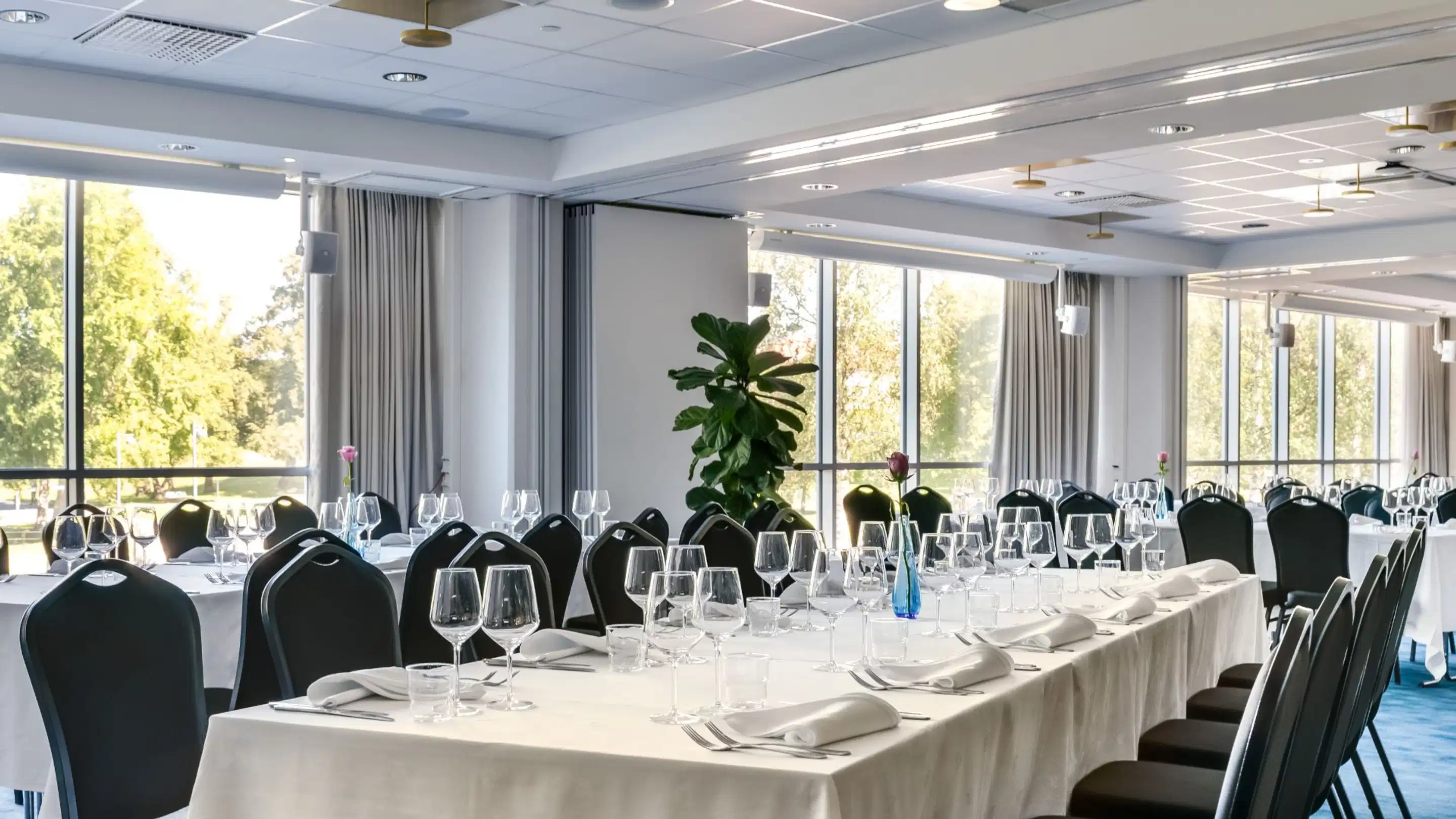 Bright banquet room set for dining with white tablecloths, glassware and cutlery. Large windows overlook greenery. Level floor surface.