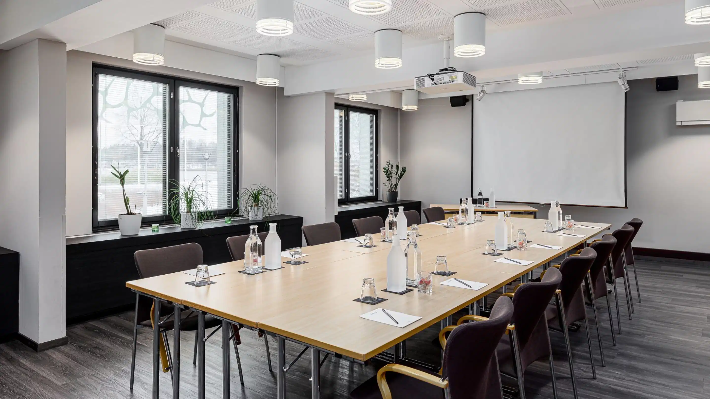 Modern meeting room with a long wooden table, water carafes and glass bottles. The space is bright with windows, even lighting and a projector.