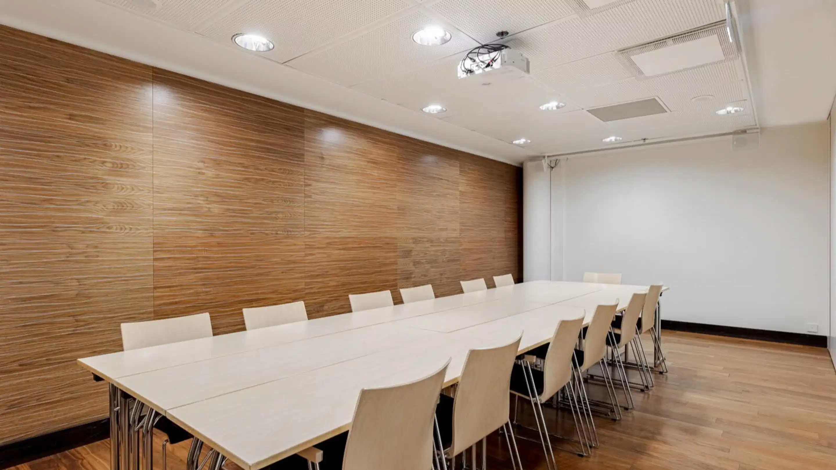 Modern conference room with long white table, light-colored chairs and wood-paneled wall. Space has even wooden flooring and ceiling-mounted projector.
