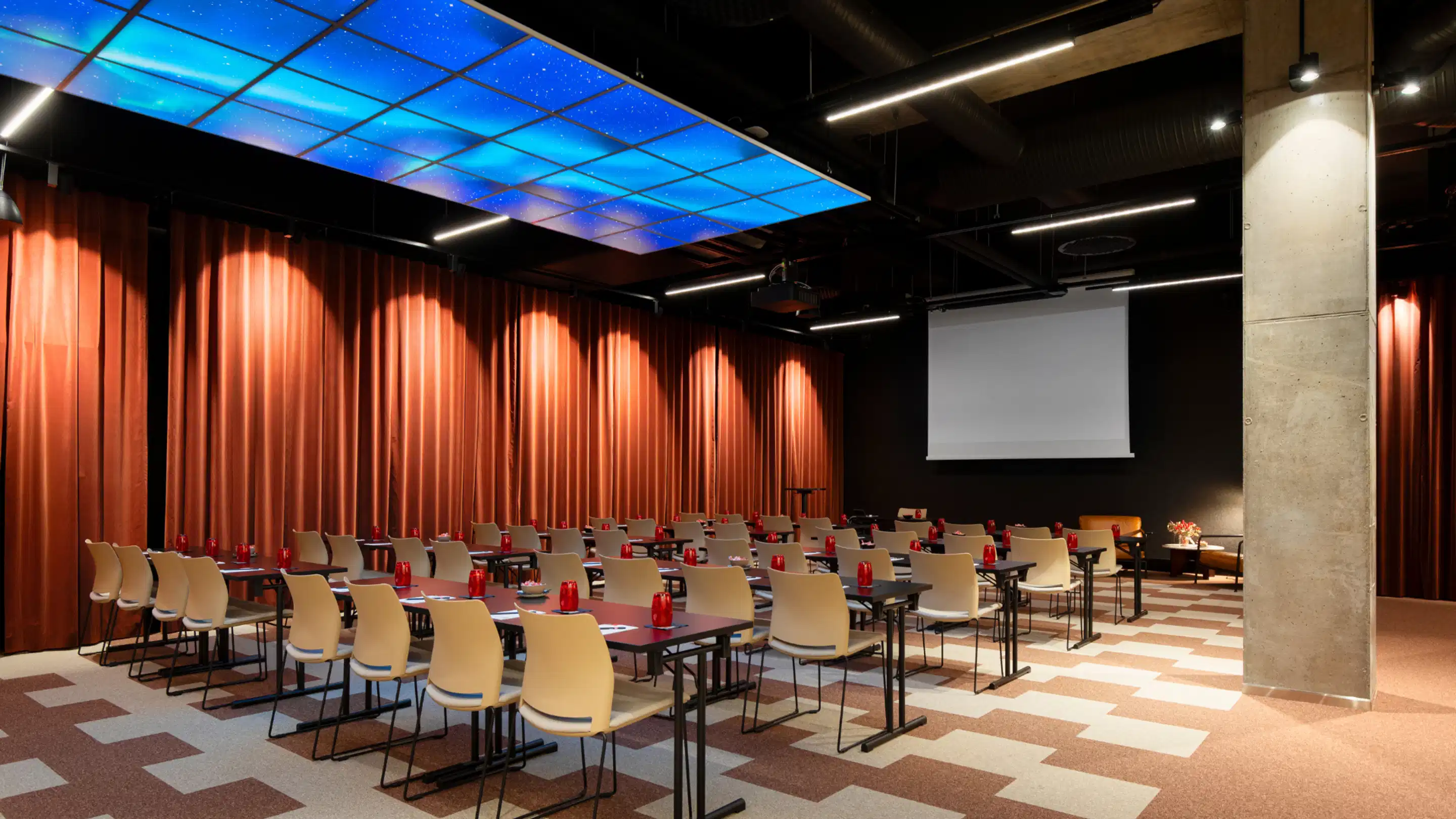 Modern auditorium with red curtains, white chairs and a blue ceiling depicting a starry sky. The floor has geometric patterns and a projection screen at the front.