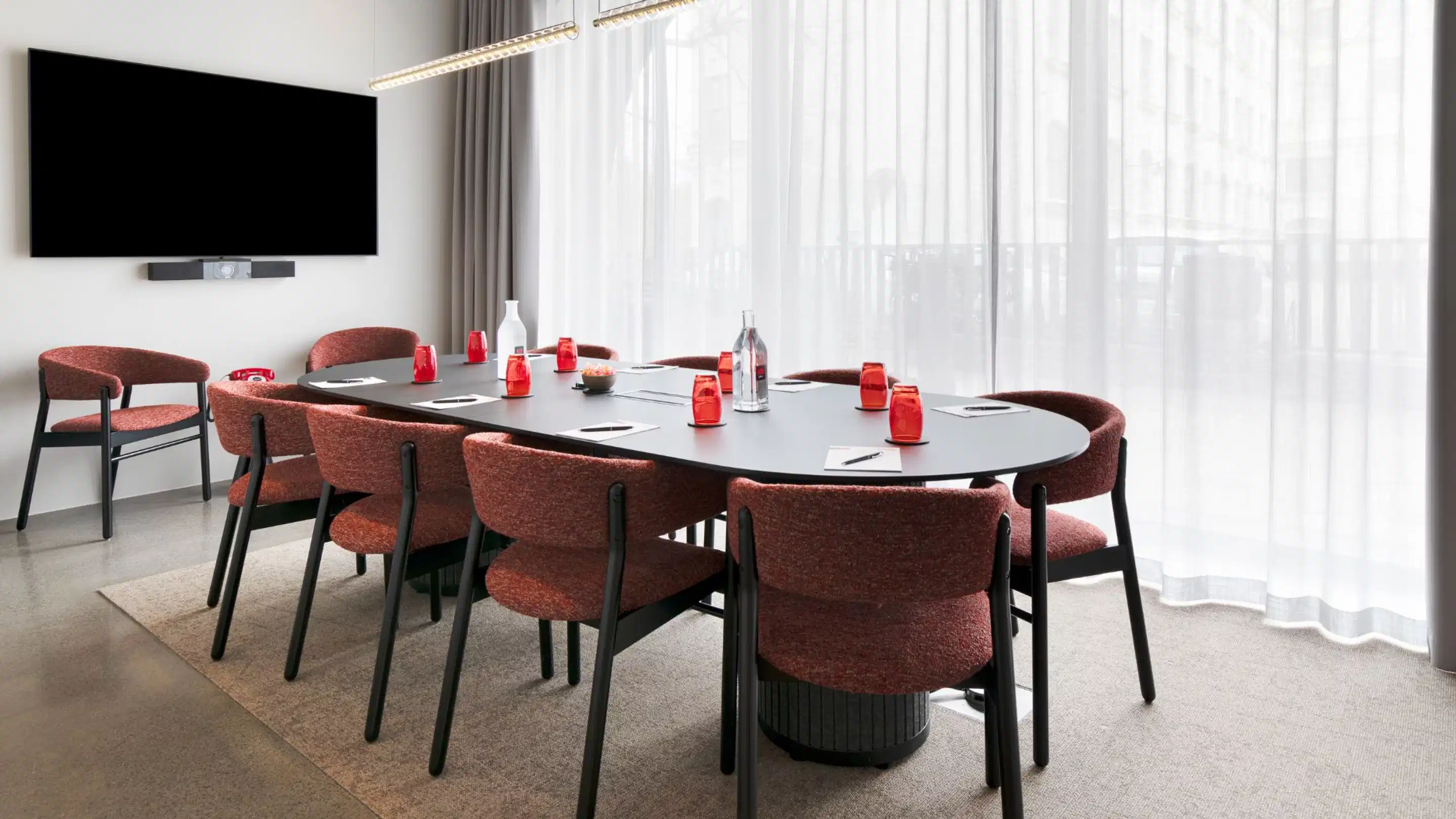 Modern meeting room with oval black table and reddish-brown chairs. Wall-mounted flat screen, large windows, and level carpeted floor.