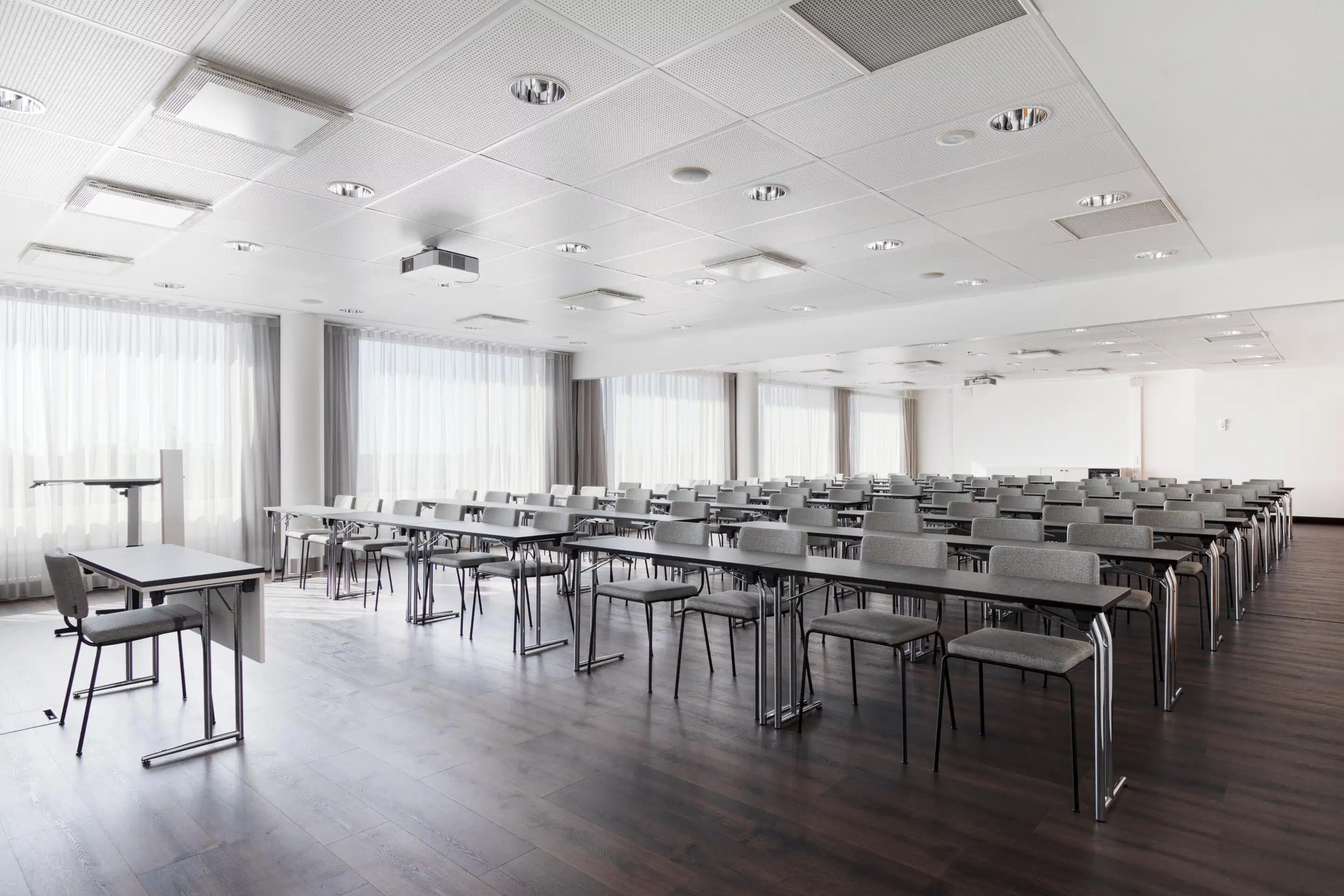 Modern conference room with long tables and gray chairs arranged in rows. The room features ceiling lights and white curtains on the walls.