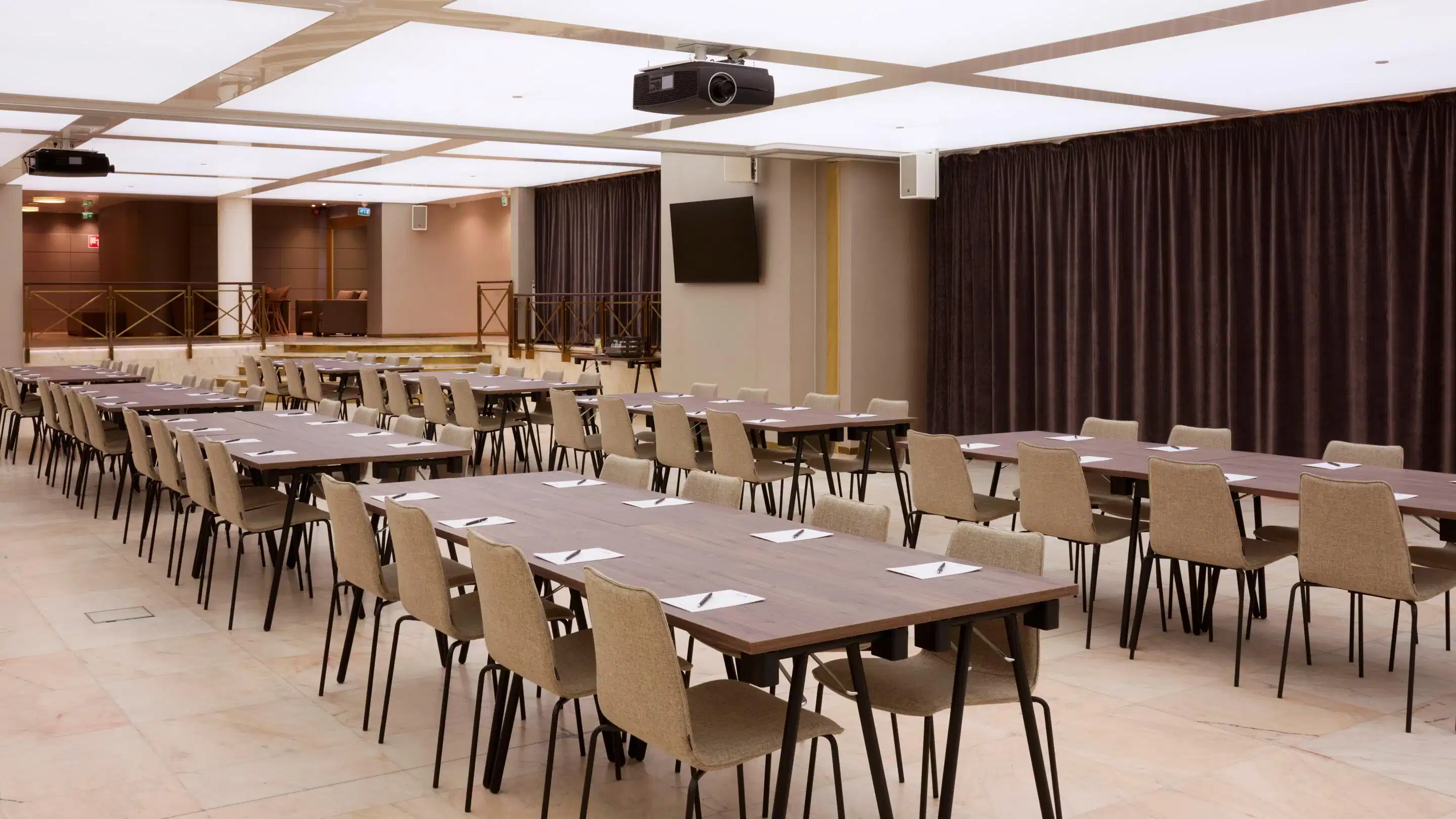 Konferenslokal med långa bord och stolar i rader, projektorer monterade i taket. Utrymmet har plant golv och ljus belysning.