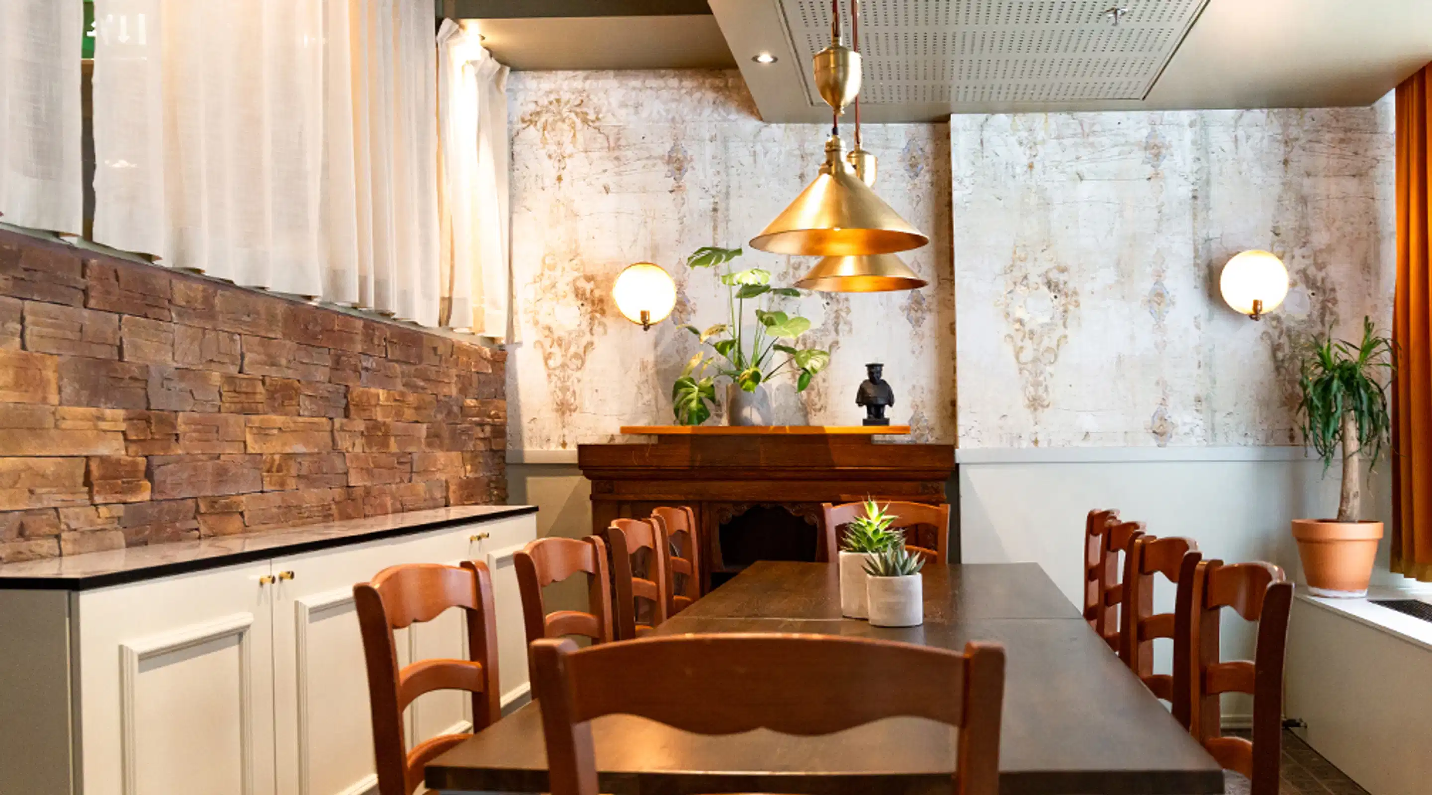 Restaurant interior with a long wooden table, chairs, stone wall, golden pendant lights and green plants. Space features wall sconces and an antique wooden cabinet.