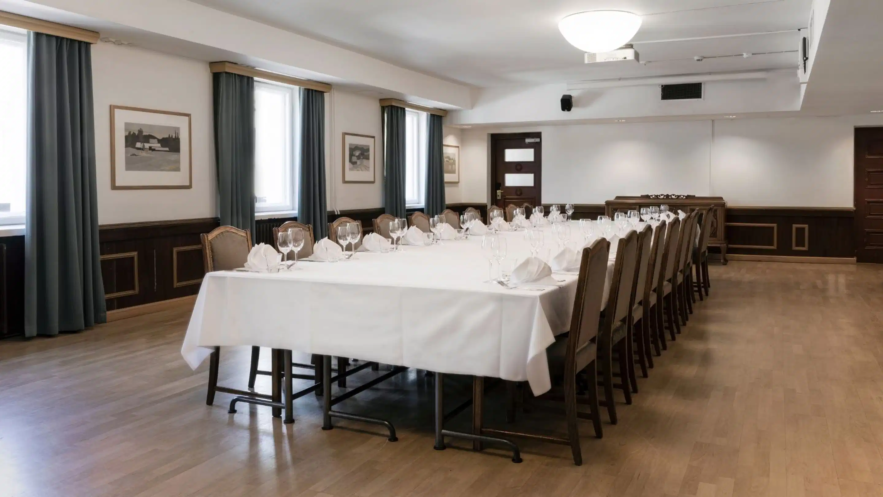 Elegantti kokoushuone, jossa pitkä pöytä katettu valkoisella liinalla ja laseilla. Tilassa puulattia, tummat verhot ja tasainen valaistus.