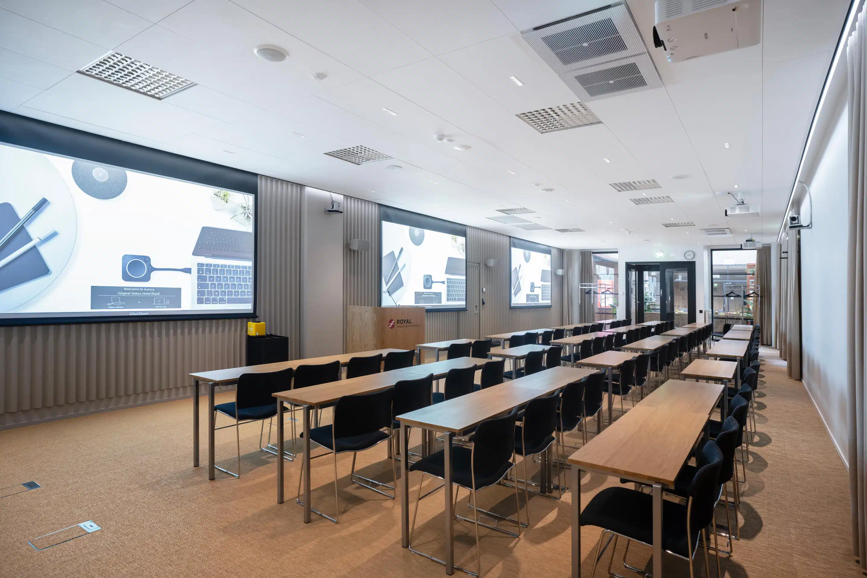 Modern classroom with long tables and chairs arranged in rows, large screens on walls, level floor surface and good lighting.