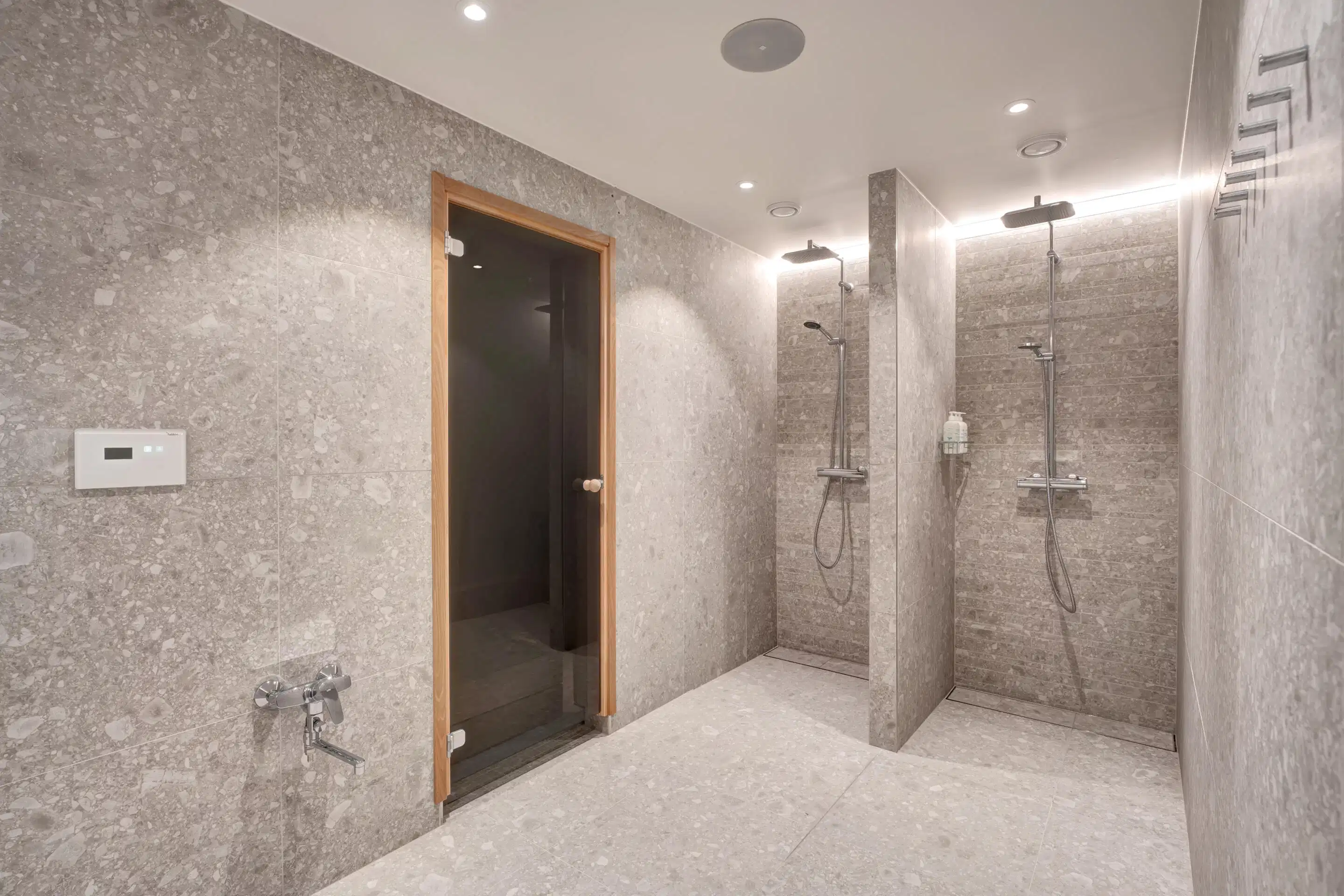 Modern bathroom with gray stone tiles featuring two showers and wooden-framed sauna door. Level floor and good lighting.