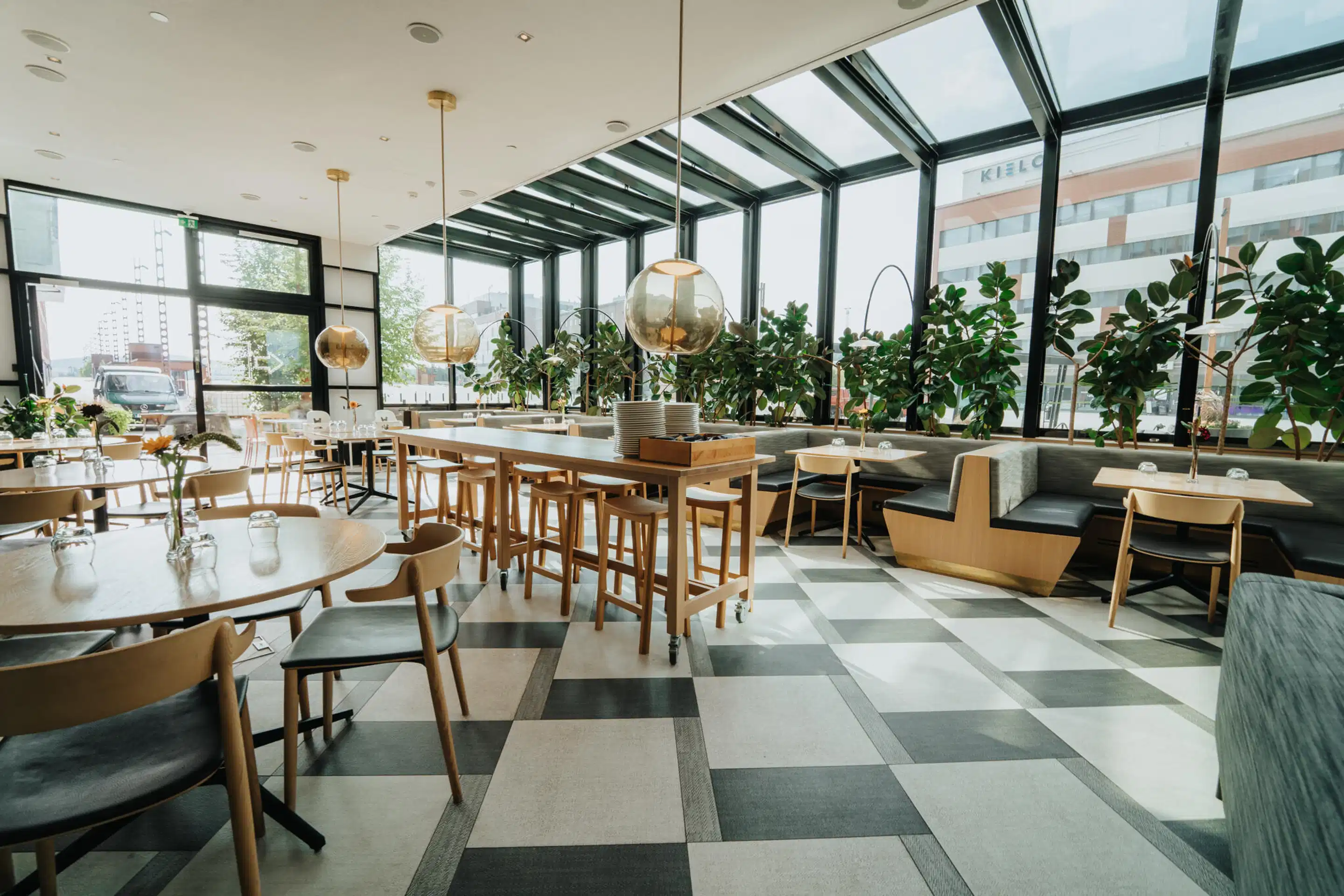 Valge restoran suurte akendega, puidust lauad ja toolid, rohelised taimed ning tasane põrand. Laelambid ripuvad kõrgel.