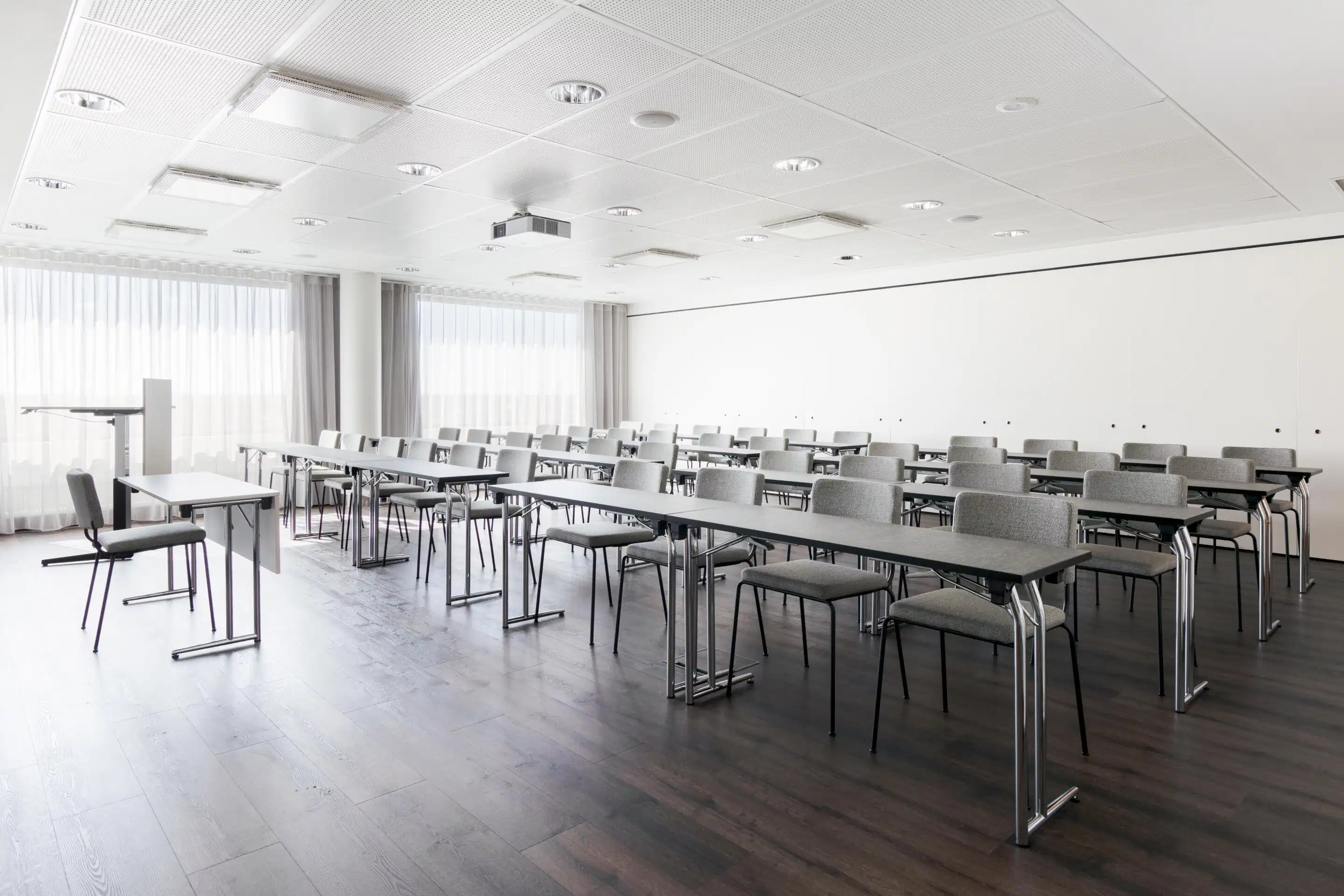 Modern, bright conference room with long tables and gray chairs arranged in rows. The room has a flat wooden floor and good ceiling lighting.