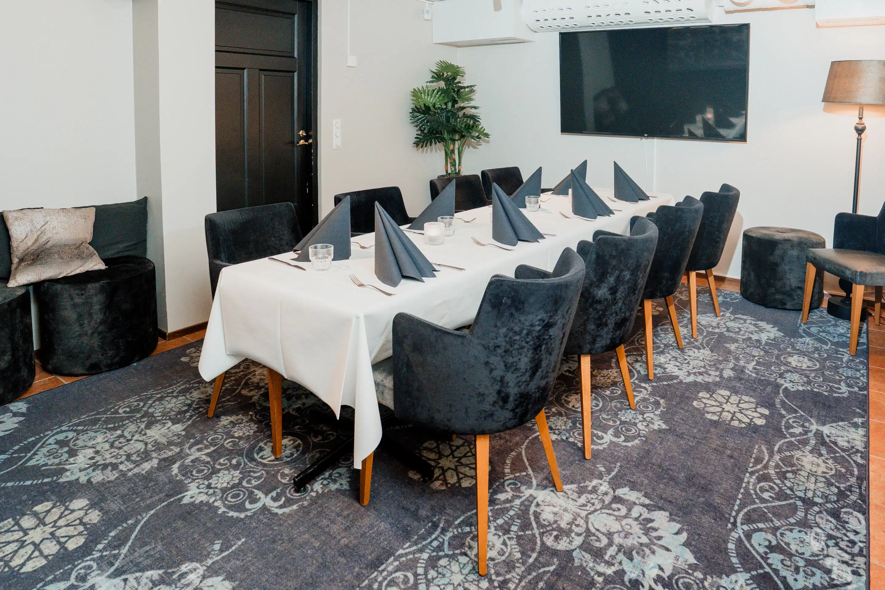Meeting room with long table covered in white cloth, blue napkins, and dark blue chairs. Wall-mounted TV, potted plant in corner, and doorway.