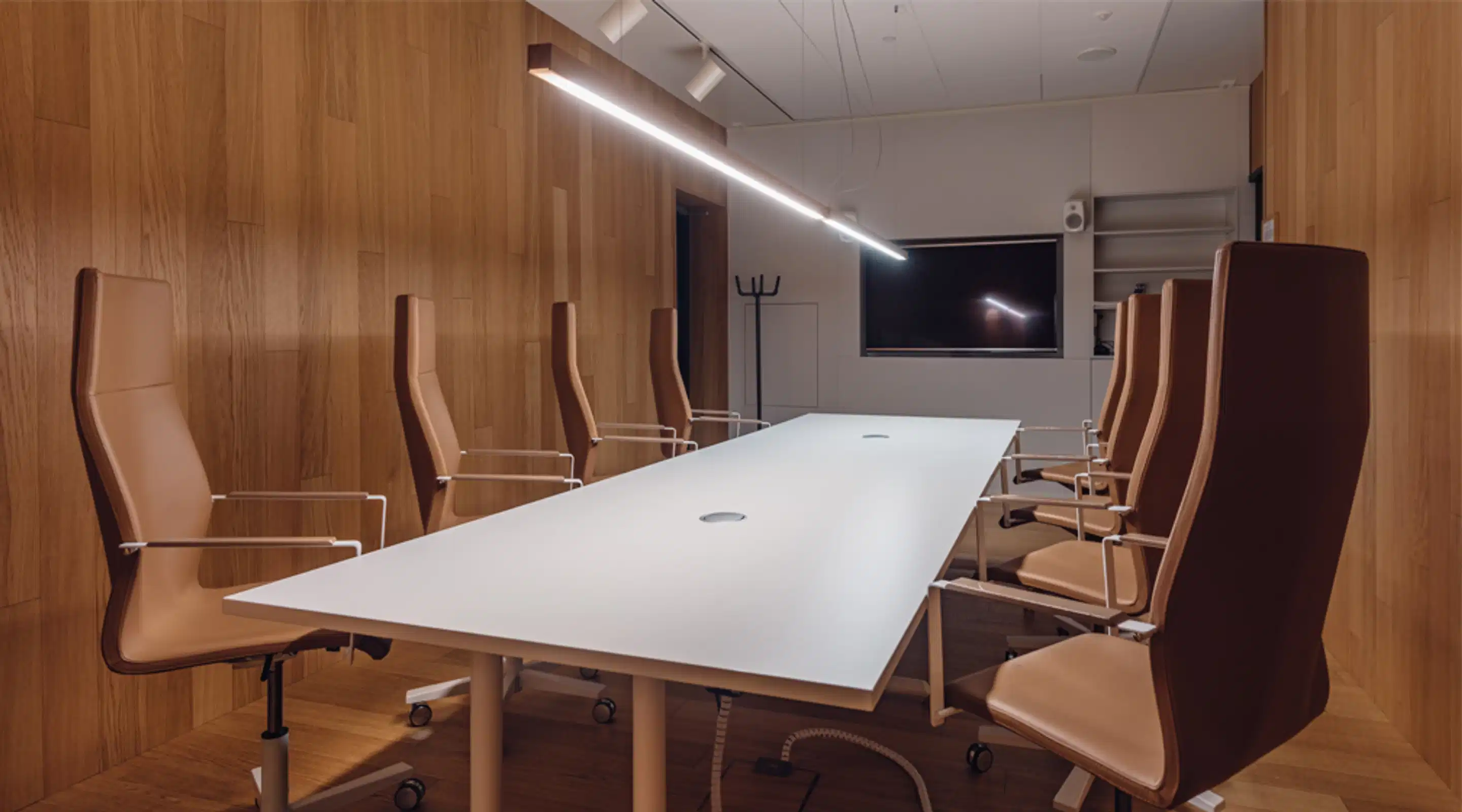 Modern meeting room with wooden paneled walls, featuring a long white table and leather office chairs. Screen on the wall and ceiling lighting.