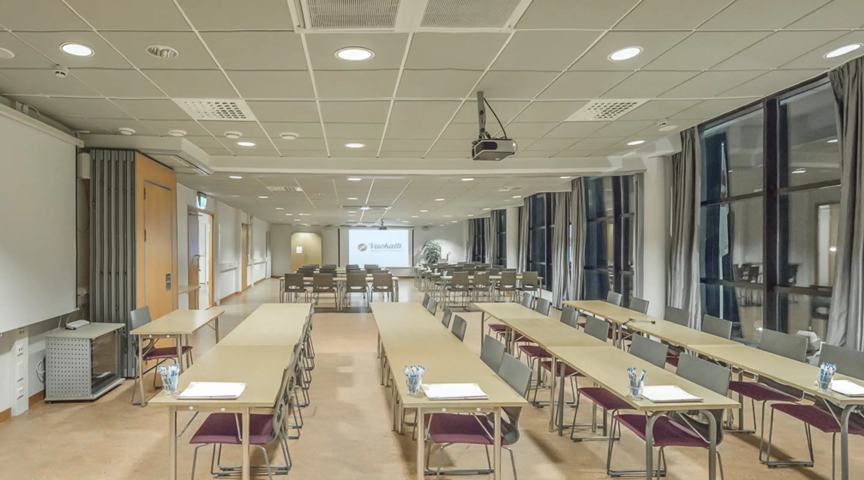 Valge koosolekuruum pikkade laudadega klassiruumi vormis, projektoriekraan ees ja suured aknad külgseinal. Ruum on avar ja tasase põrandaga.