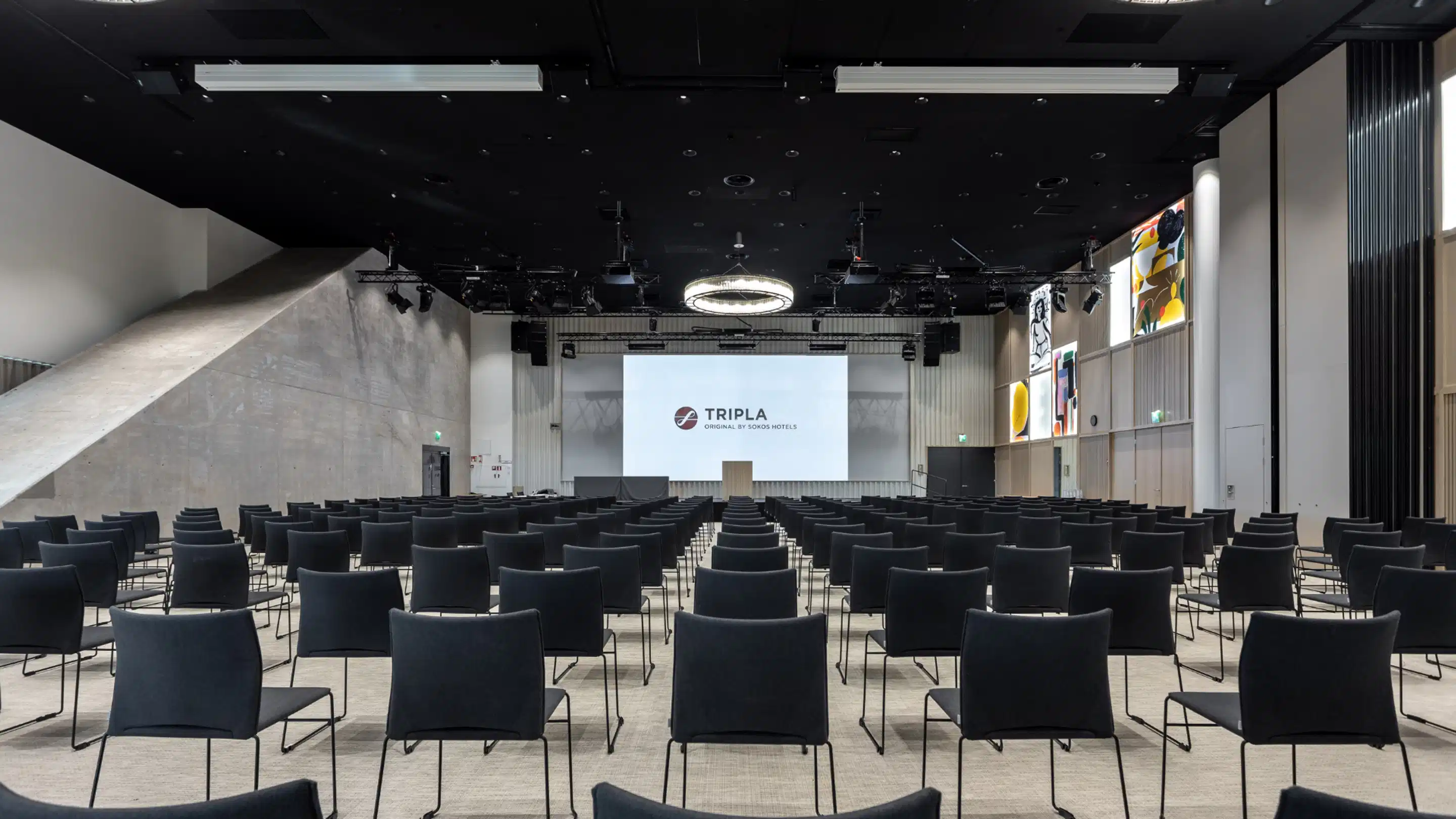 Moderni auditorio, jossa on rivit sinisiä tuoleja, lavalla valkoinen projektorikangas, korkeat betoniset seinät ja musta kattopinta.