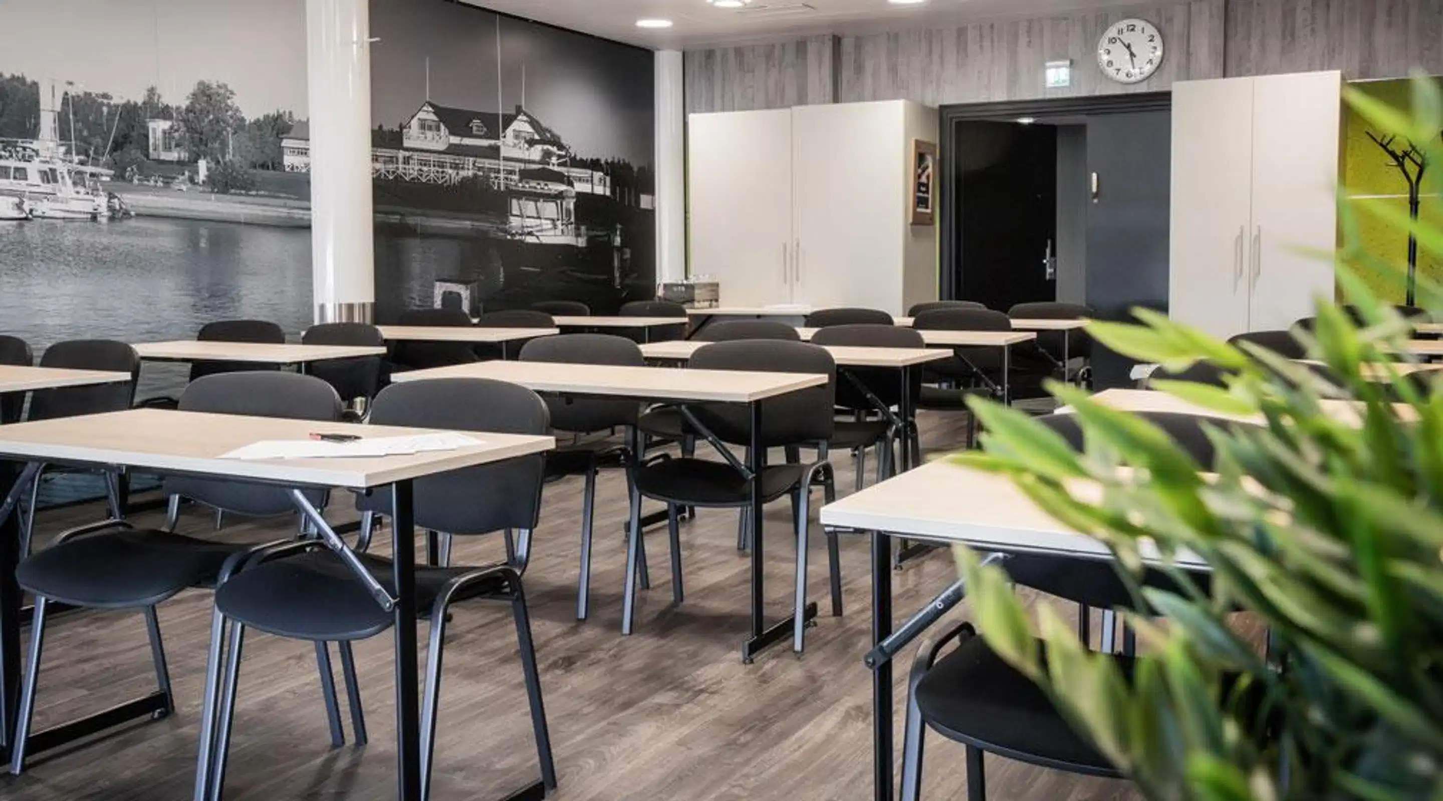 Koulutustila, jossa on vaaleita pöytiä ja mustia tuoleja. Taustalla on valokuvatapetti satamasta ja tiivis, kova lattiapinta.