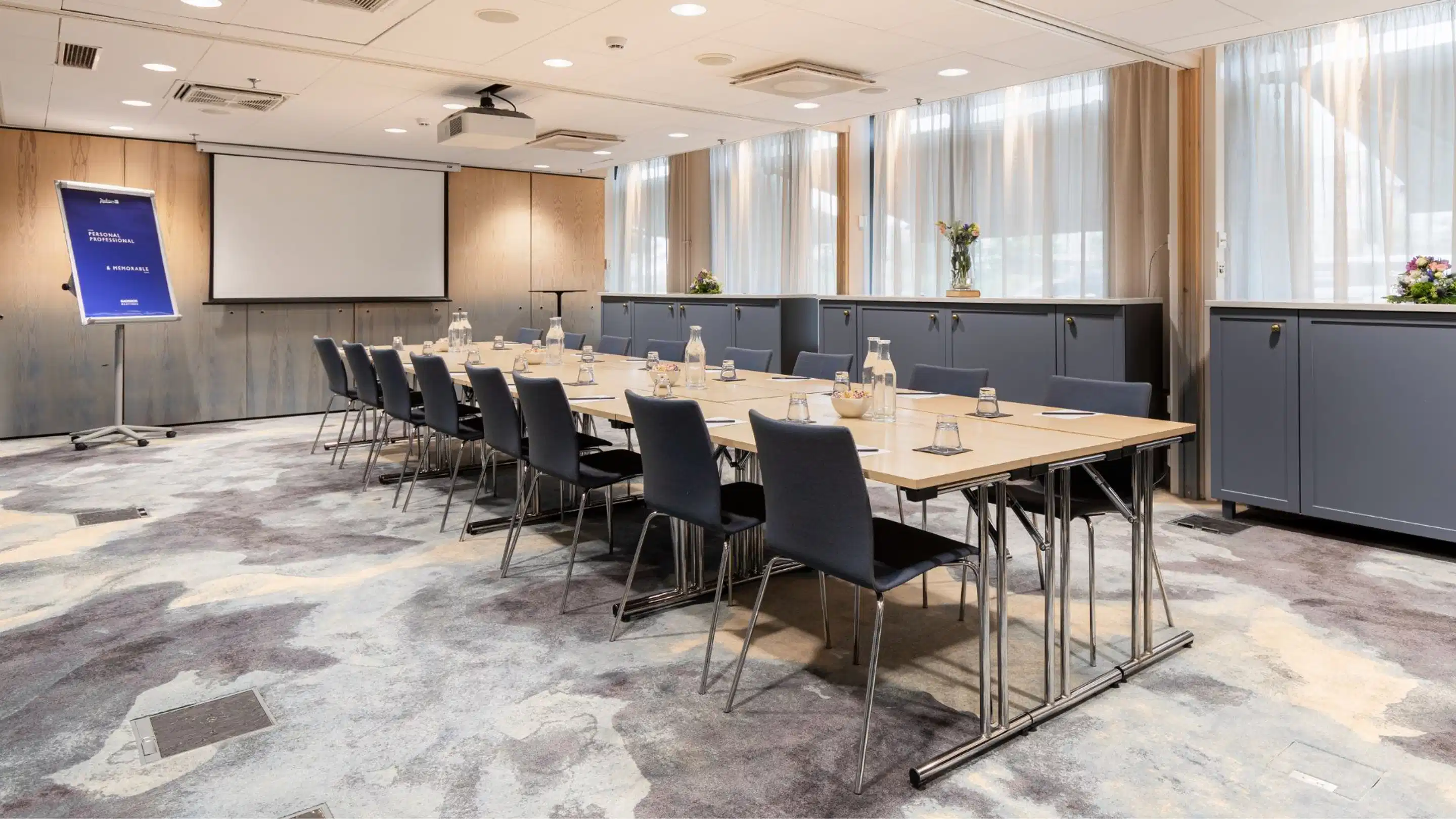 Conference room with long wooden table surrounded by dark grey chairs, projection screen and ceiling projector. Large windows, wooden wall panels and marbled patterned floor.