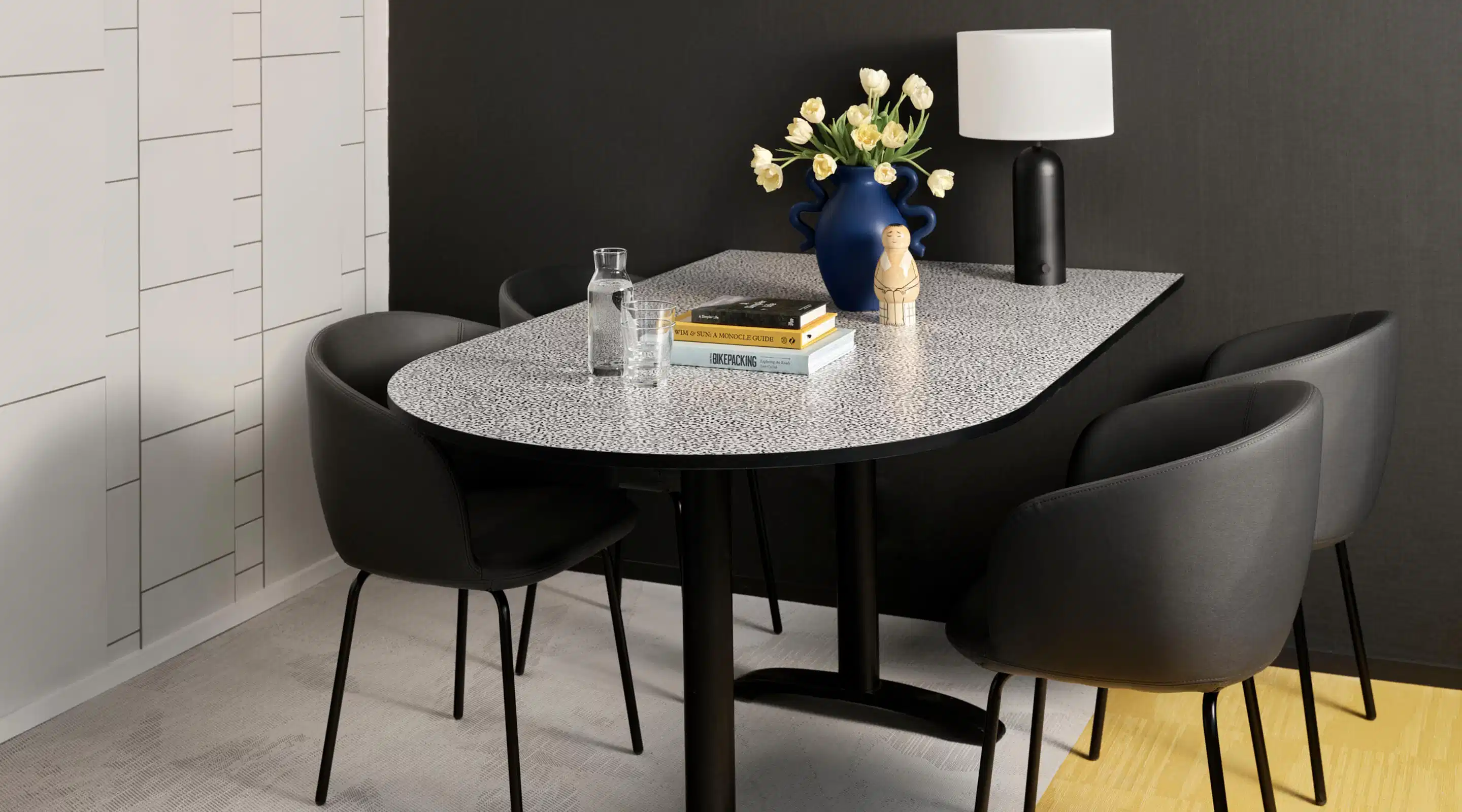 Modern dining area with terrazzo table, four black chairs, blue vase with light tulips and black table lamp with light shade.
