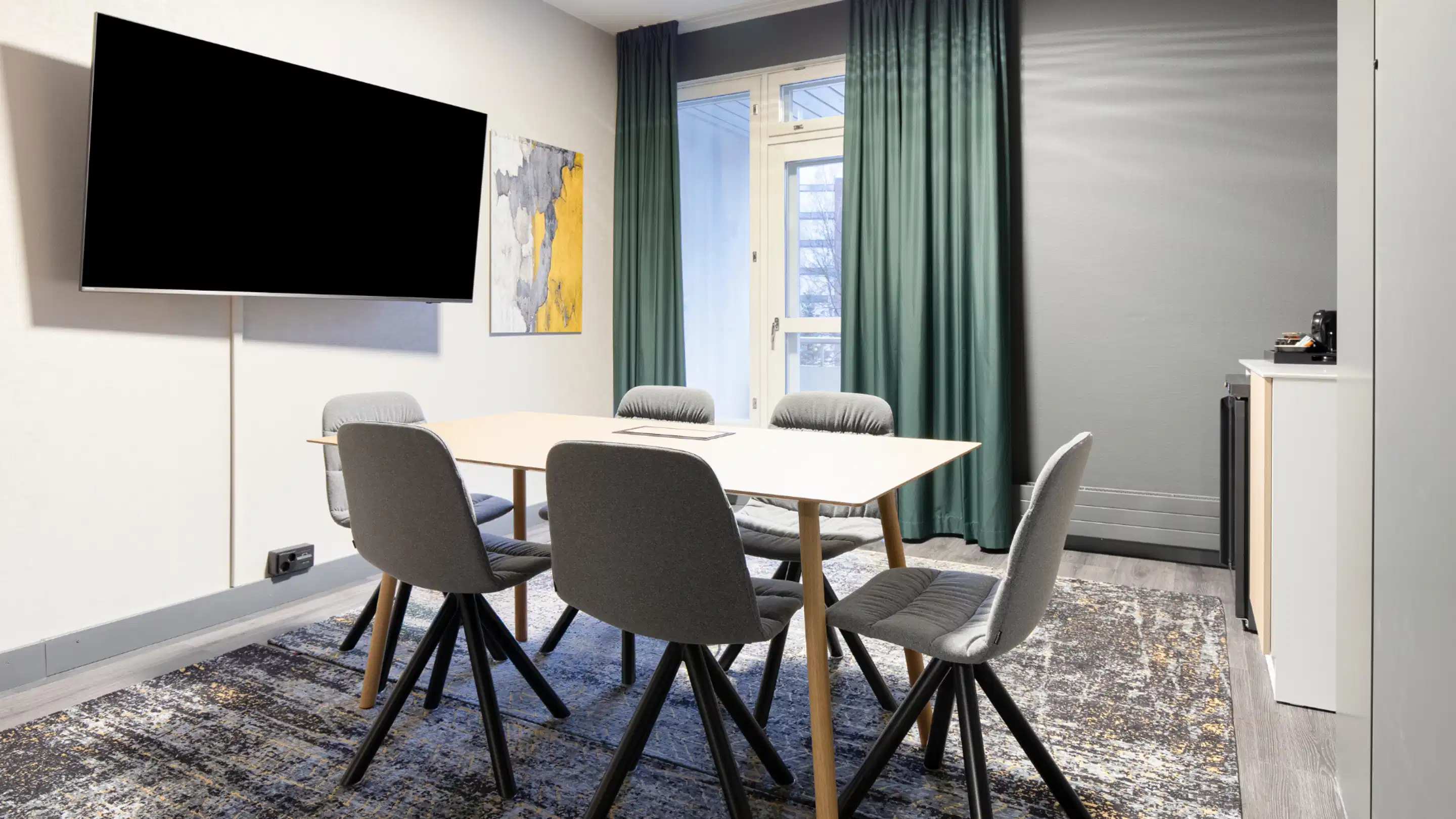 Modern meeting room with wooden table, six grey chairs, wall-mounted large TV and green curtains. The floor is hard with a patterned area rug, natural light coming through the window.