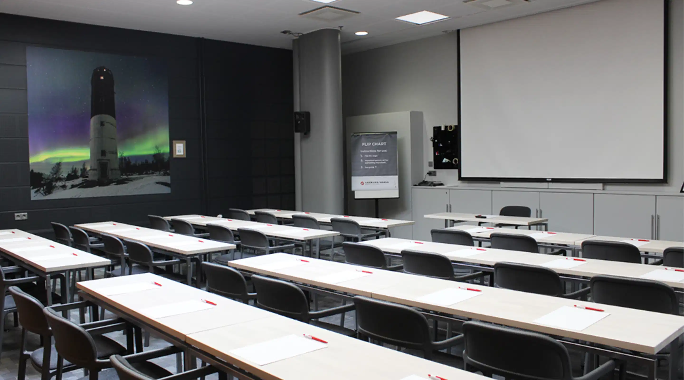Classroom with long tables and chairs. Large image of a lighthouse under northern lights on the wall, projection screen at the front.