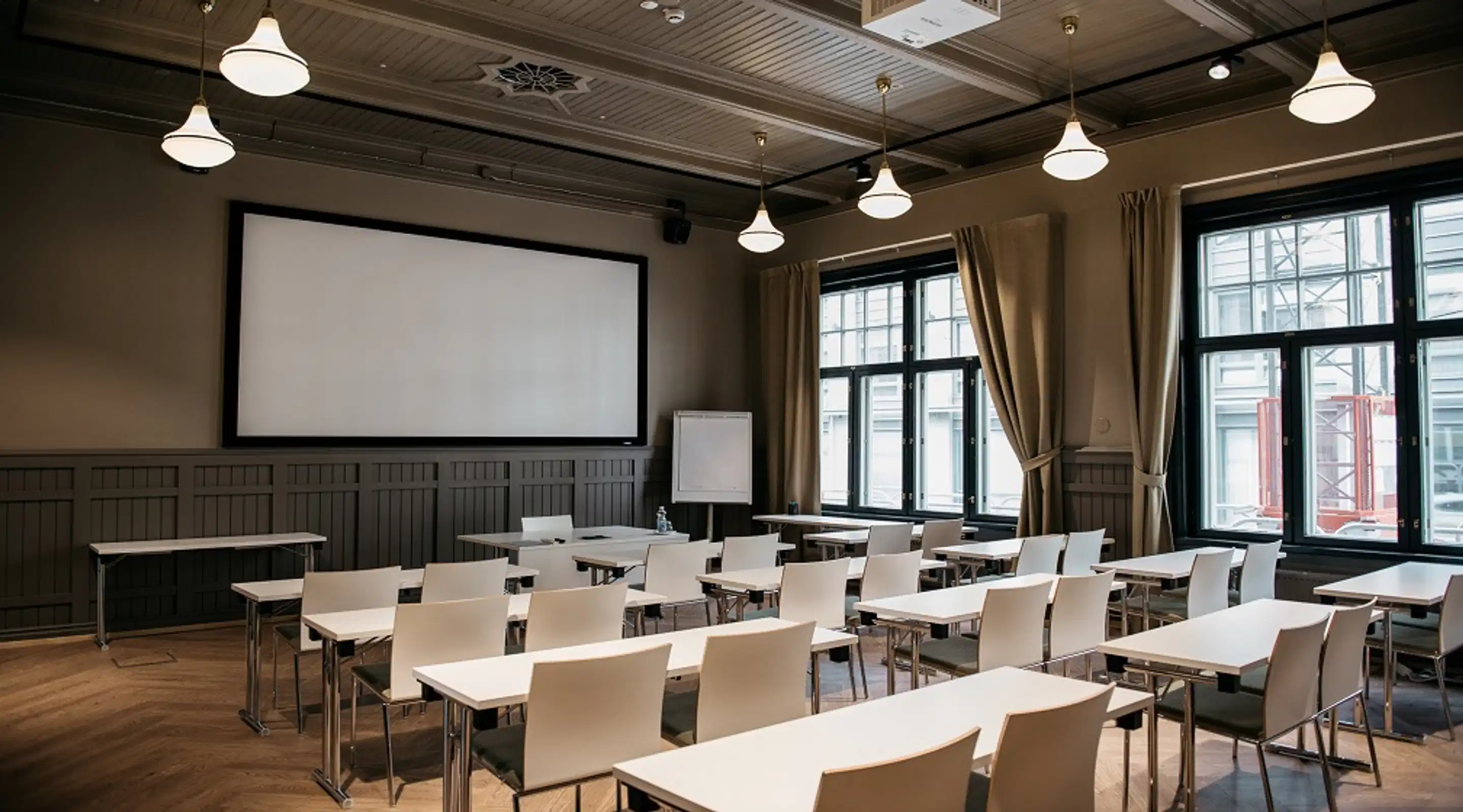 Classroom-style meeting room with white tables and chairs. Projection screen at front, large windows with beige curtains.