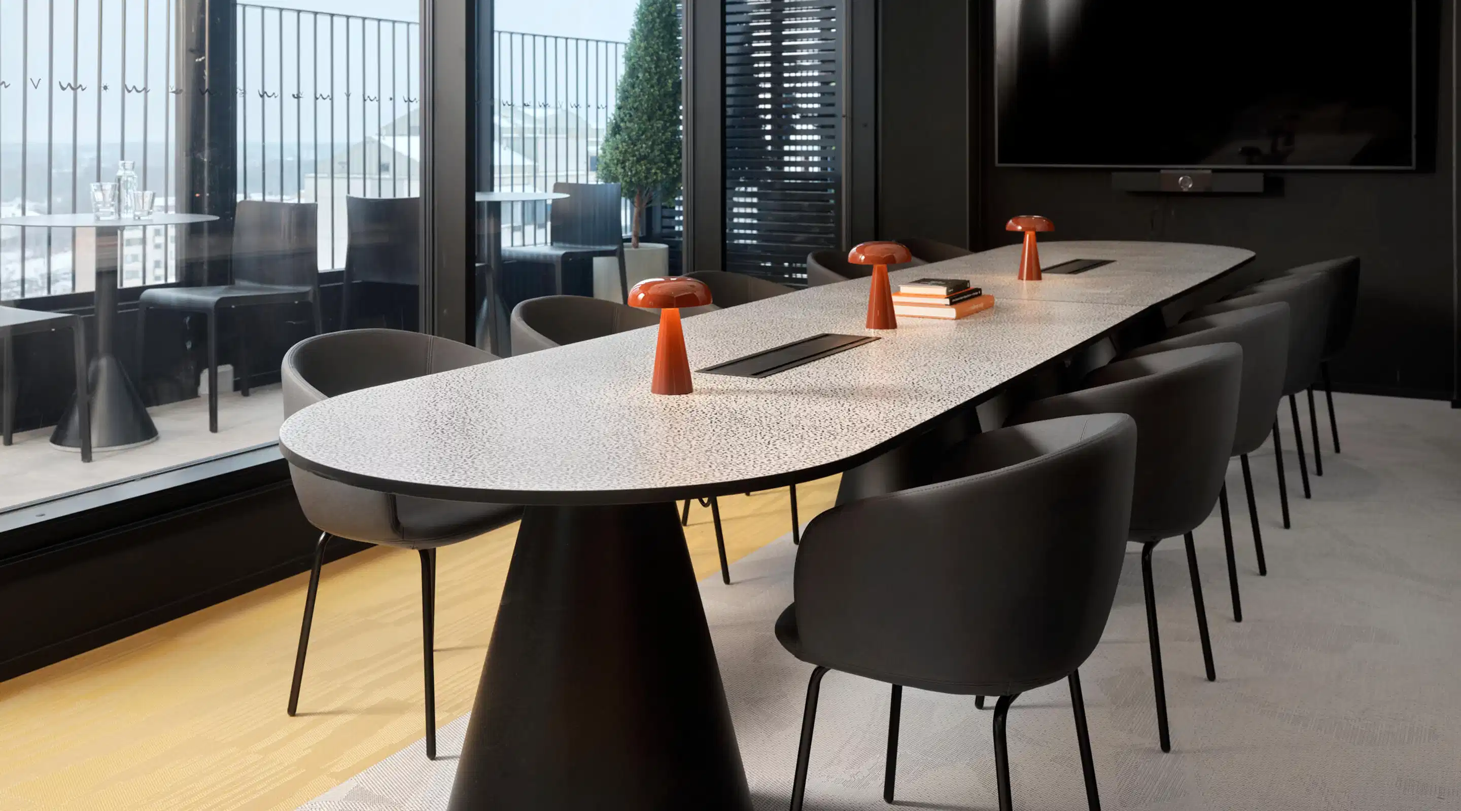 Modern conference room with white oval table and black chairs. Orange table lamps and large windows allowing natural light. Wooden floor and wall-mounted TV screen.